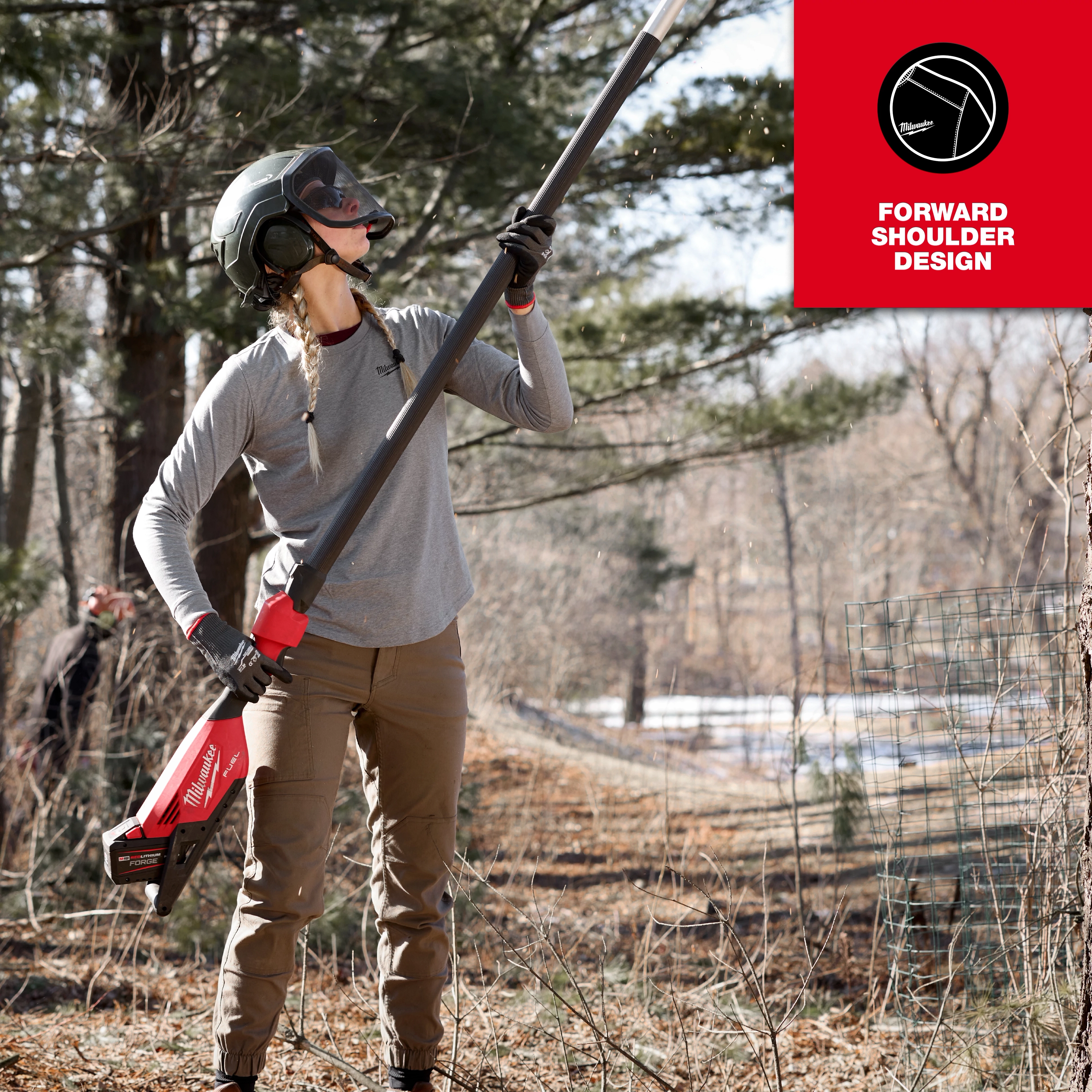 A woman wearing the Women's FREEFLEX™ Hybrid Tee - Long Sleeve is working in a wooded area using a power tool. She is wearing safety gear including a helmet and gloves. The tee features a forward shoulder design.