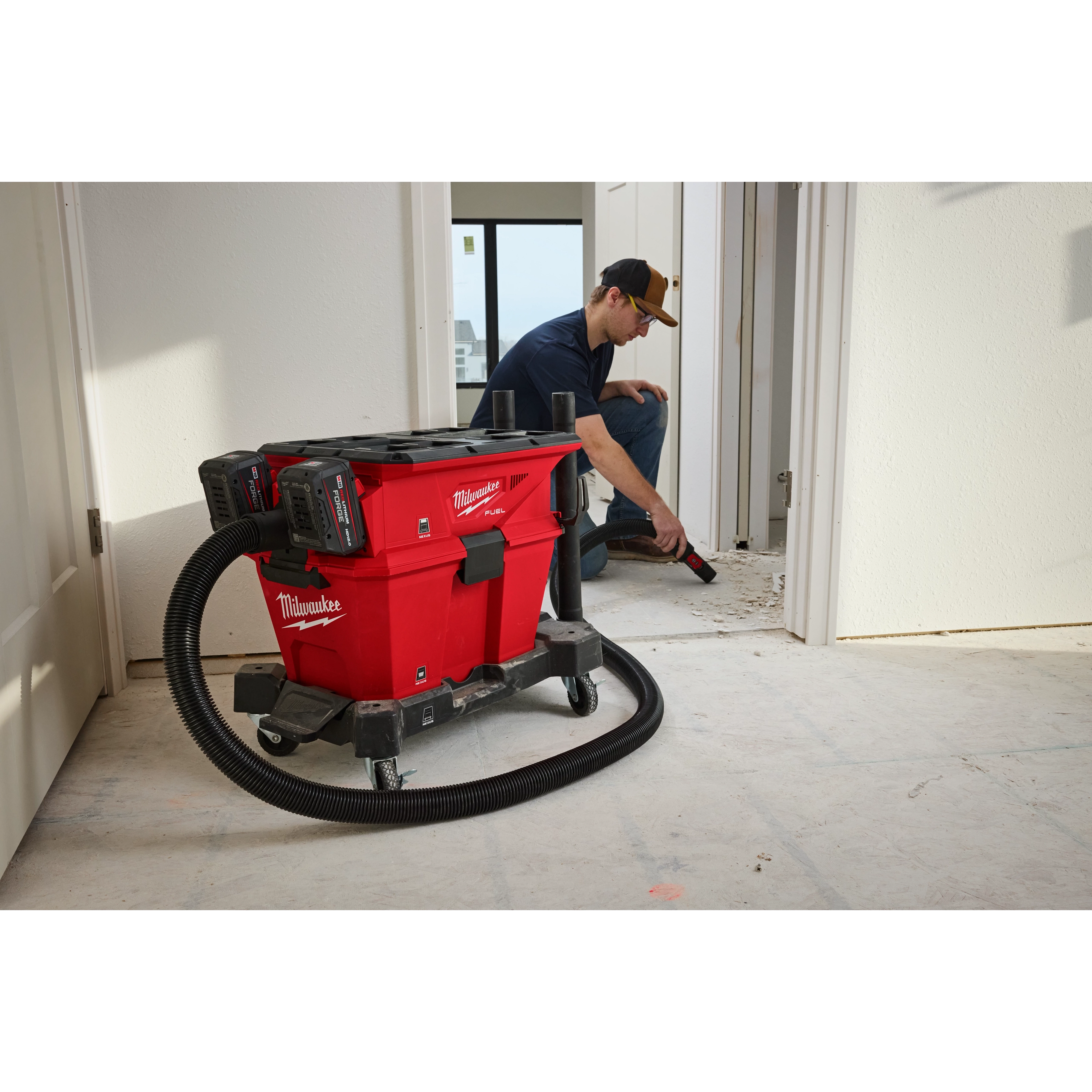 The M18 FUEL™ NEXUS™ Dual Battery Wet/Dry Vacuum Motor Head with PACKOUT™ Compatibility & VACLINK™ is shown in use on a job site. The vacuum is red with black accents and connected to a flexible hose, efficiently cleaning debris from the floor.
