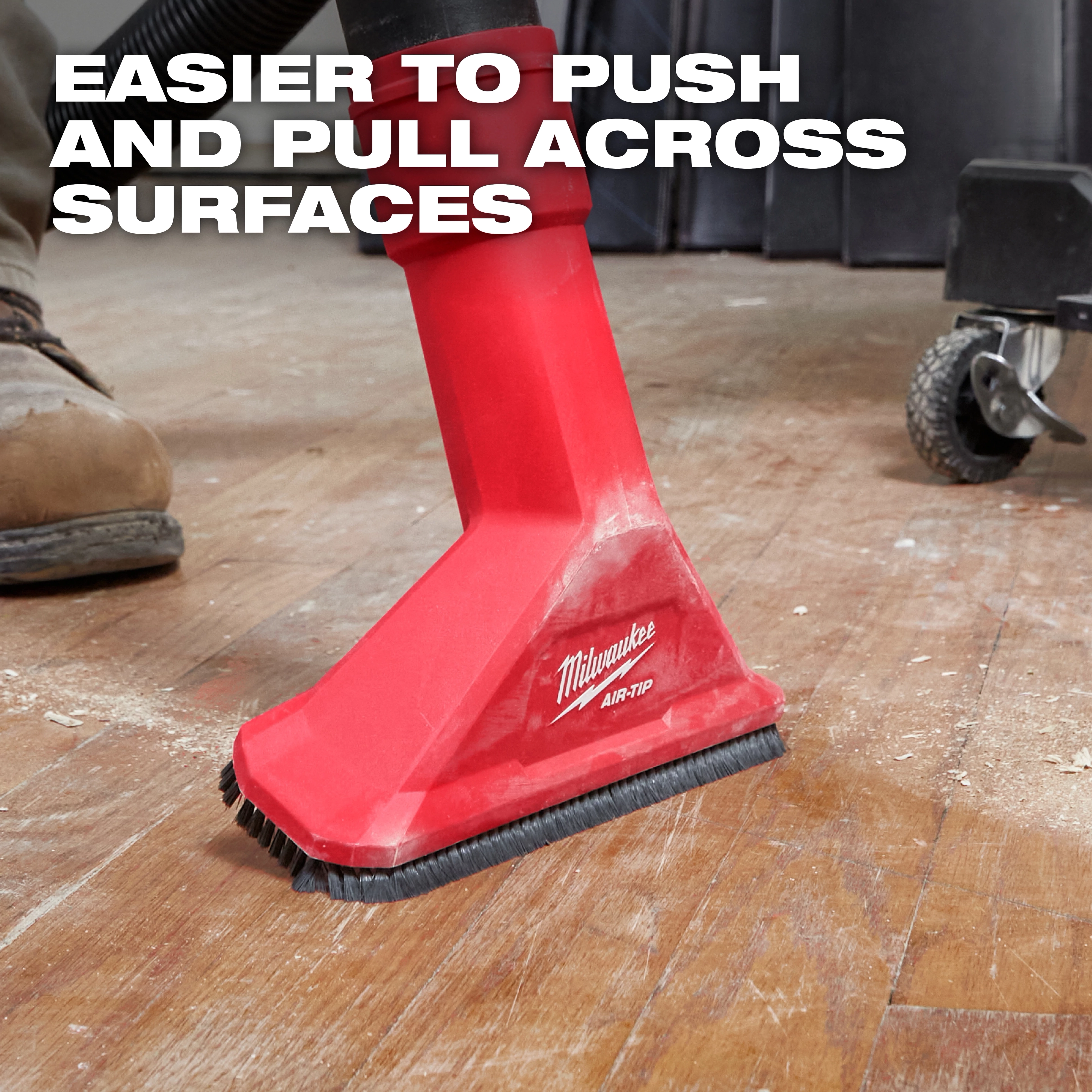 The image shows a red Milwaukee AIR-TIP™ 2-1/2" Rocking Utility Nozzle with Brushes being used on a wooden floor. The nozzle is attached to a vacuum and has brushes at its base. Text on the image reads, "Easier to push and pull across surfaces."
