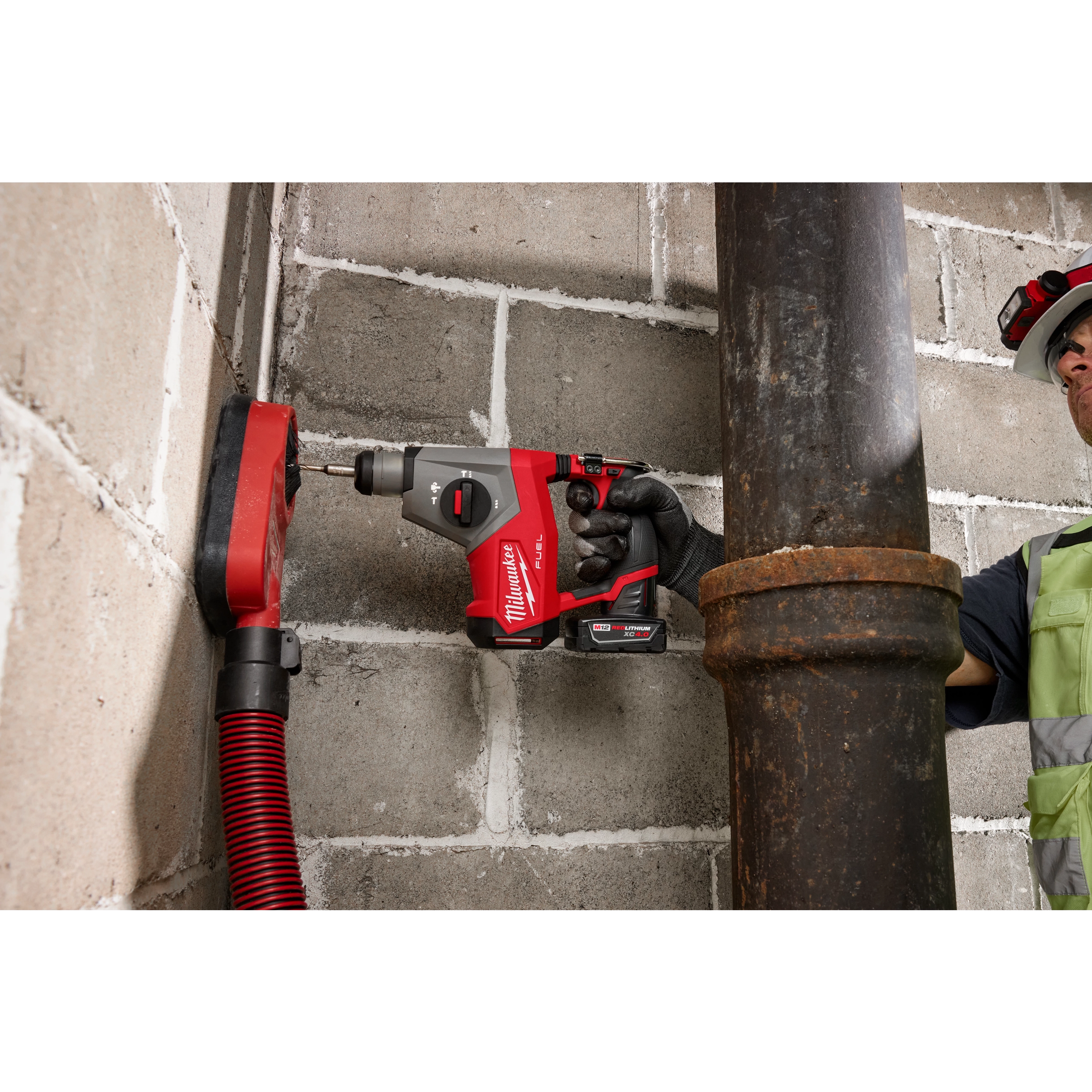 A person using an M12 FUEL™ 5/8” SDS Plus Rotary Hammer to drill into a concrete wall next to a large metal pipe. The person is wearing safety gear, including a hard hat and high-visibility vest. A red hose is connected to the drilling area for debris extraction.