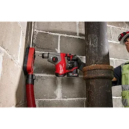 A person using an M12 FUEL™ 5/8” SDS Plus Rotary Hammer to drill into a concrete wall next to a large metal pipe. The person is wearing safety gear, including a hard hat and high-visibility vest. A red hose is connected to the drilling area for debris extraction.