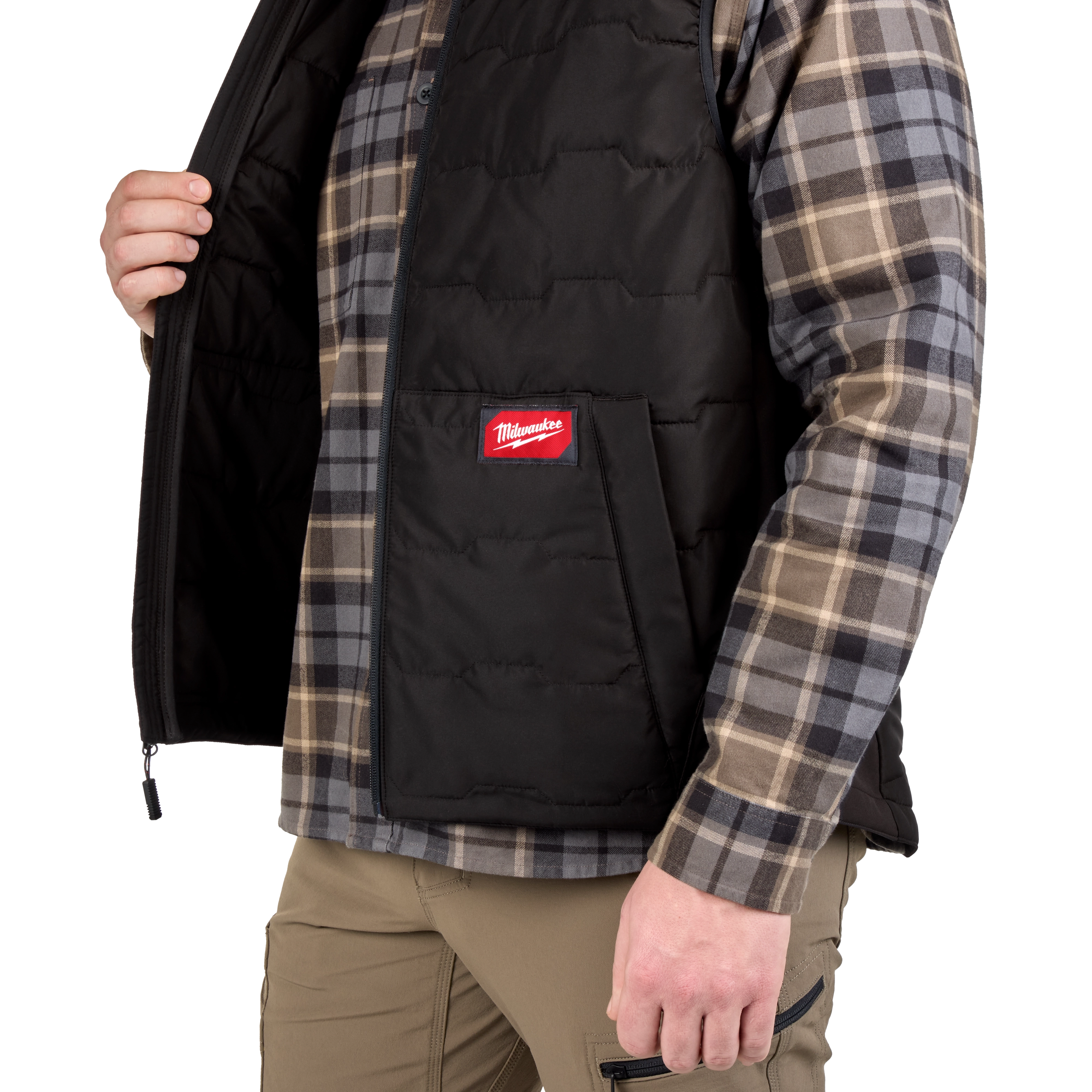 A person wears a black FREEFLEX™ Lightweight Insulated Vest over a gray and beige plaid shirt. The vest has a zipper, front pockets, and a red Milwaukee logo on the left side. The person is shown from the chest down, holding the vest open with one hand.