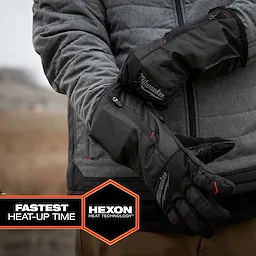 Person wearing black Milwaukee Heated Winter Gloves with zippers, standing outdoors in a gray jacket. The gloves feature Hexon heat technology and have the fastest heat-up time, as indicated by the text in the bottom left corner. Background includes blurred landscape and soft lighting.