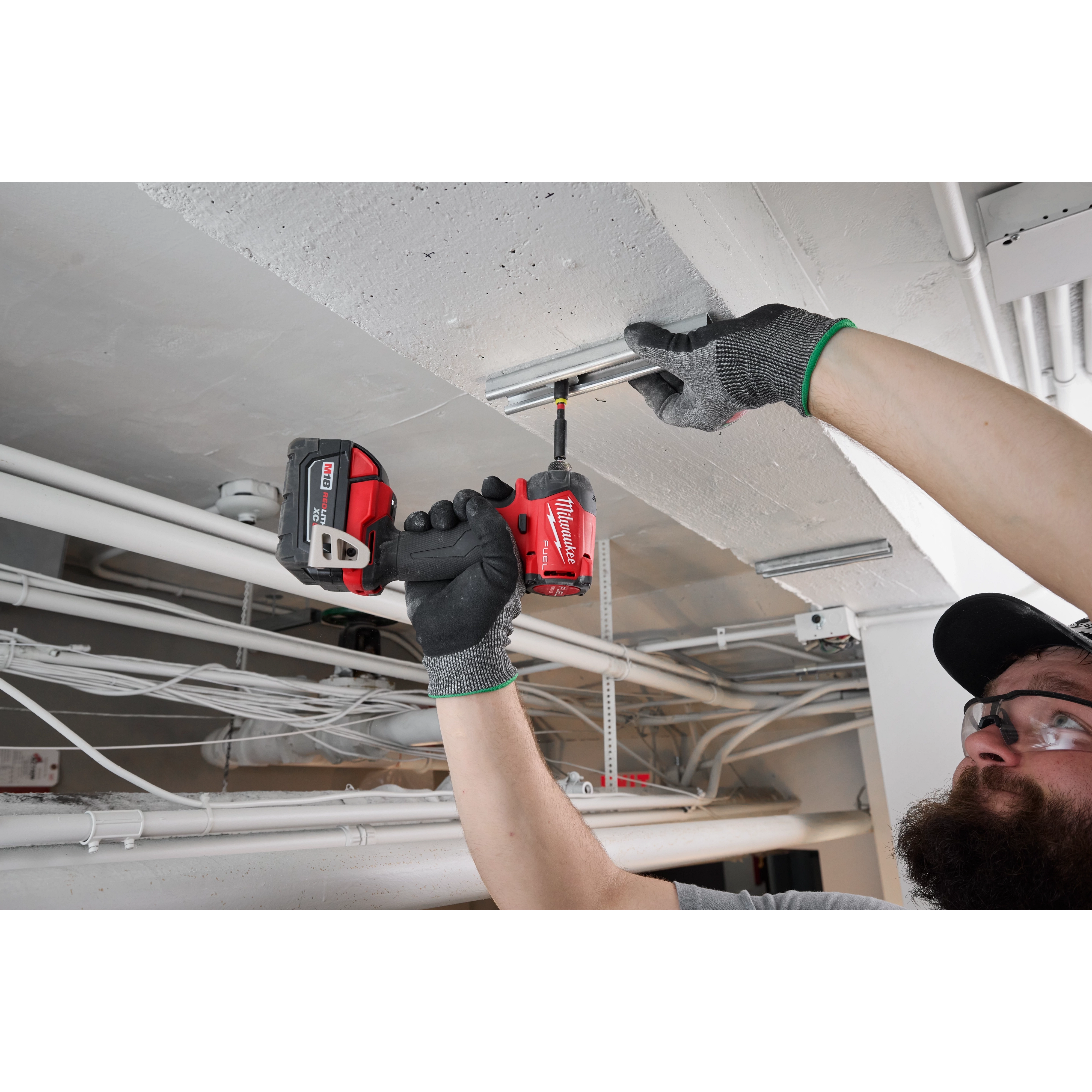 A person uses the M18 FUEL™ SURGE™ 1/4" Hex Hydraulic Driver to secure a metal bracket to a ceiling. The tool is red and black, and the user is wearing grey gloves with green trim. Various pipes and cables are visible in the background.