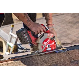 A person is using a Milwaukee circular saw equipped with a 7-1/4” 24T NITRUS™ Carbide Framing & Demolition Circular Saw Blade to cut through roofing material. The saw is generating dust, indicating active cutting. The user is wearing a red bracelet and has tattoos on their forearm.