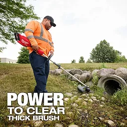 A person wearing a high-visibility orange jacket is using a Milwaukee power tool to trim grass and brush near a drainage pipe surrounded by rocks. The text "POWER TO CLEAR THICK BRUSH" is displayed in the lower left corner. Trees and a building are visible in the background.