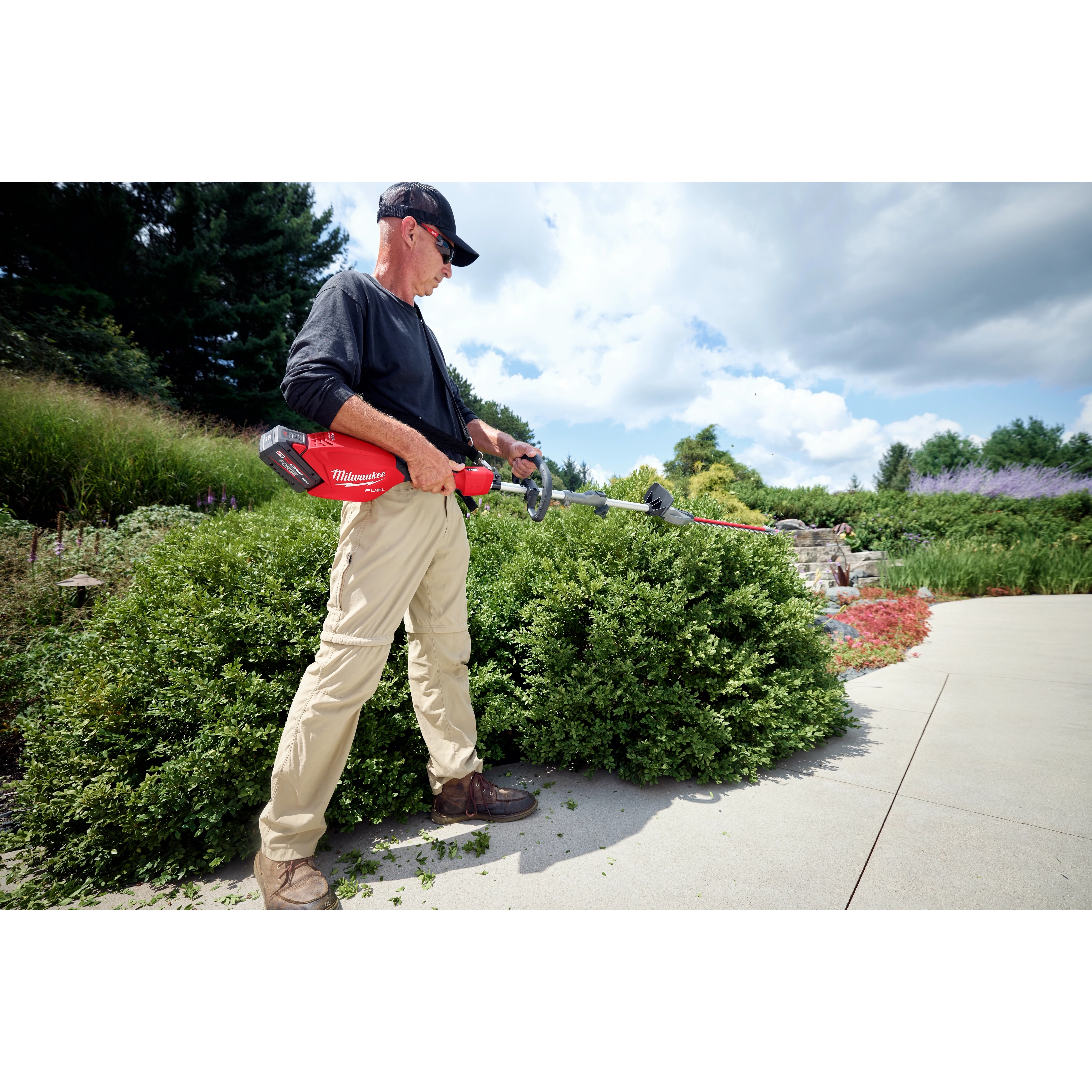 Person trimming hedges with a red Milwaukee hedge trimmer in a garden on a sunny day.