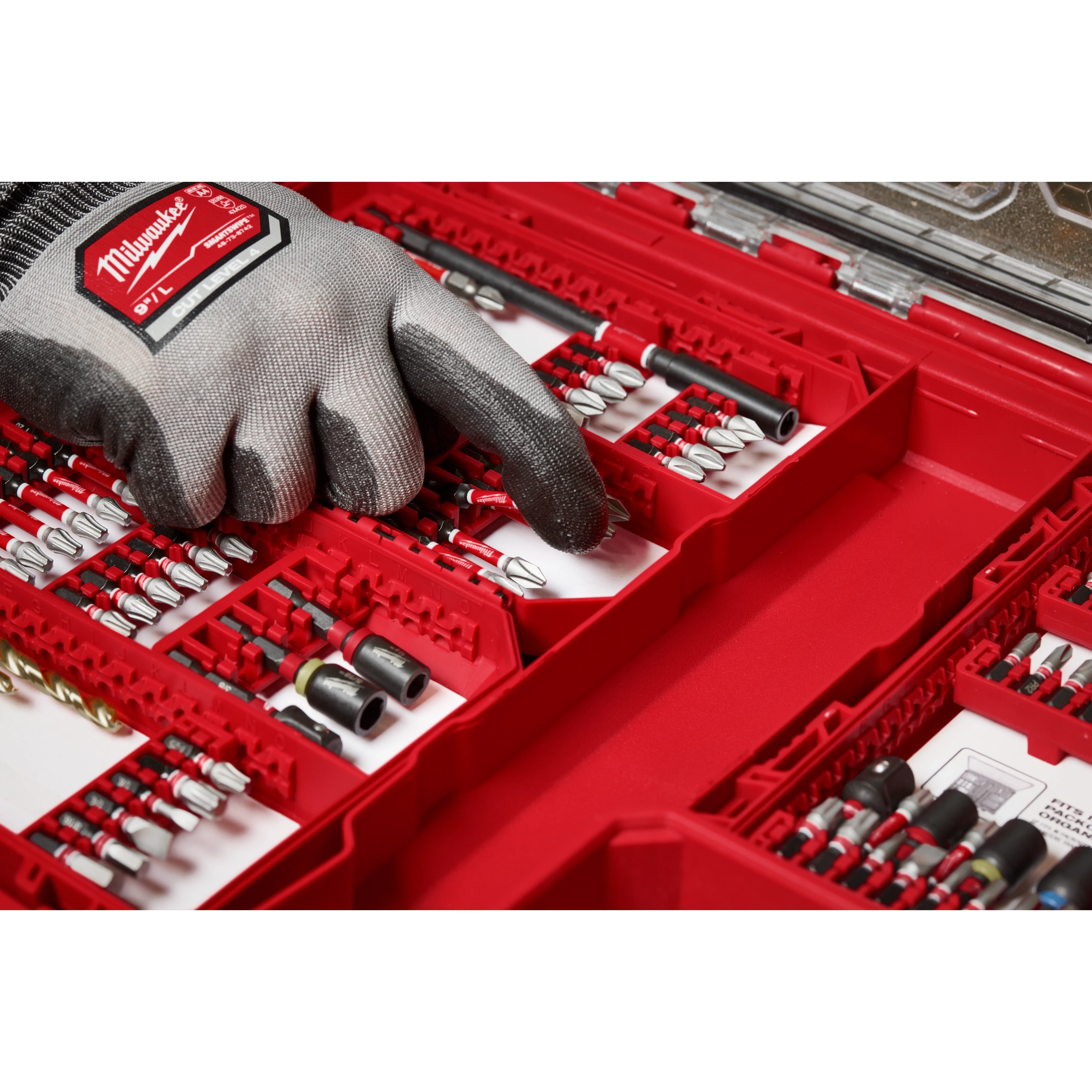 A gloved hand selecting a piece from the SHOCKWAVE Impact Duty™ Drill & Drive Set - 60PC. The red case organizes a variety of drill bits and drivers, neatly arranged in rows. The Milwaukee branding is visible on the glove.