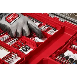 A gloved hand selecting a piece from the SHOCKWAVE Impact Duty™ Drill & Drive Set - 60PC. The red case organizes a variety of drill bits and drivers, neatly arranged in rows. The Milwaukee branding is visible on the glove.