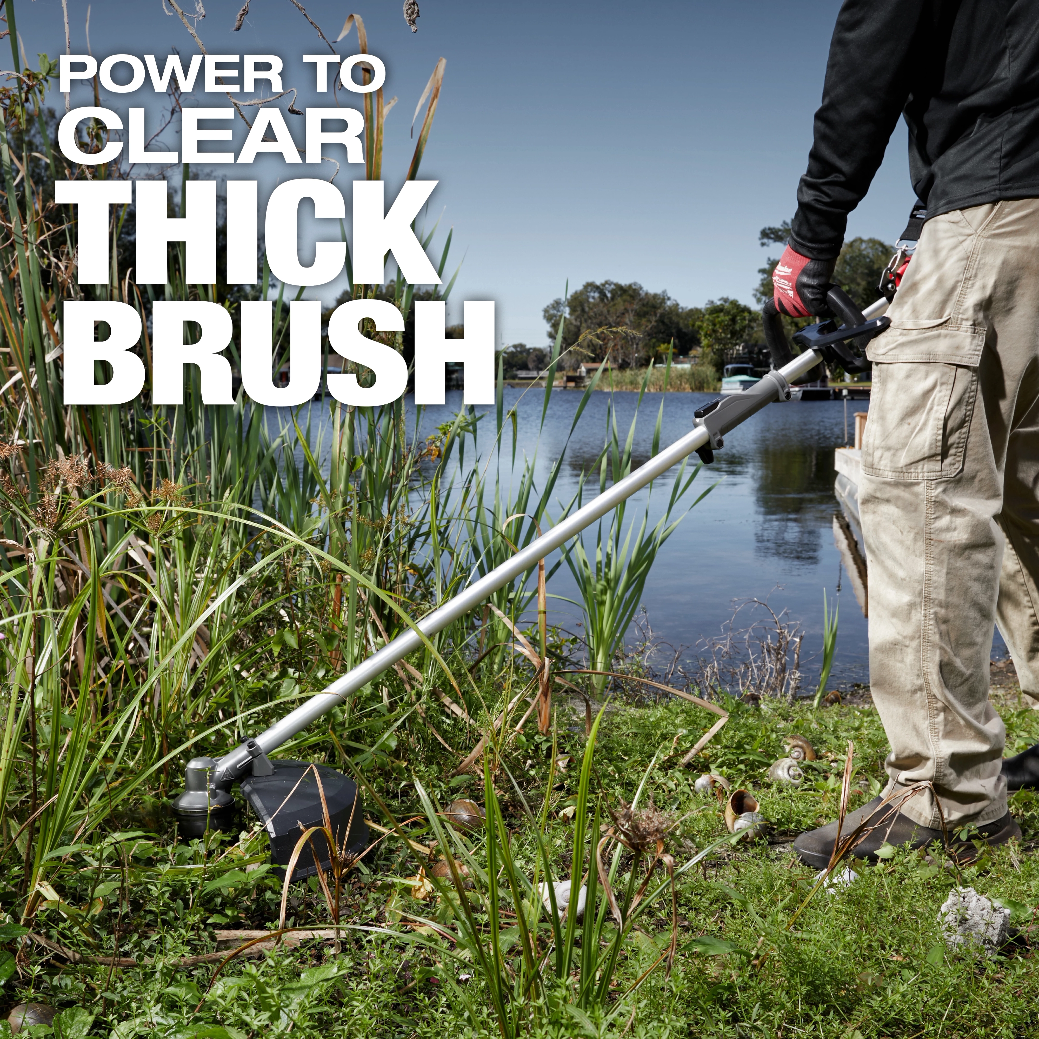 A person in work pants and gloves uses a brush cutter to clear thick vegetation by a lakeside. The text on the image reads "Power to Clear Thick Brush."