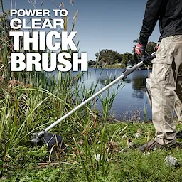 A person in work pants and gloves uses a brush cutter to clear thick vegetation by a lakeside. The text on the image reads "Power to Clear Thick Brush."