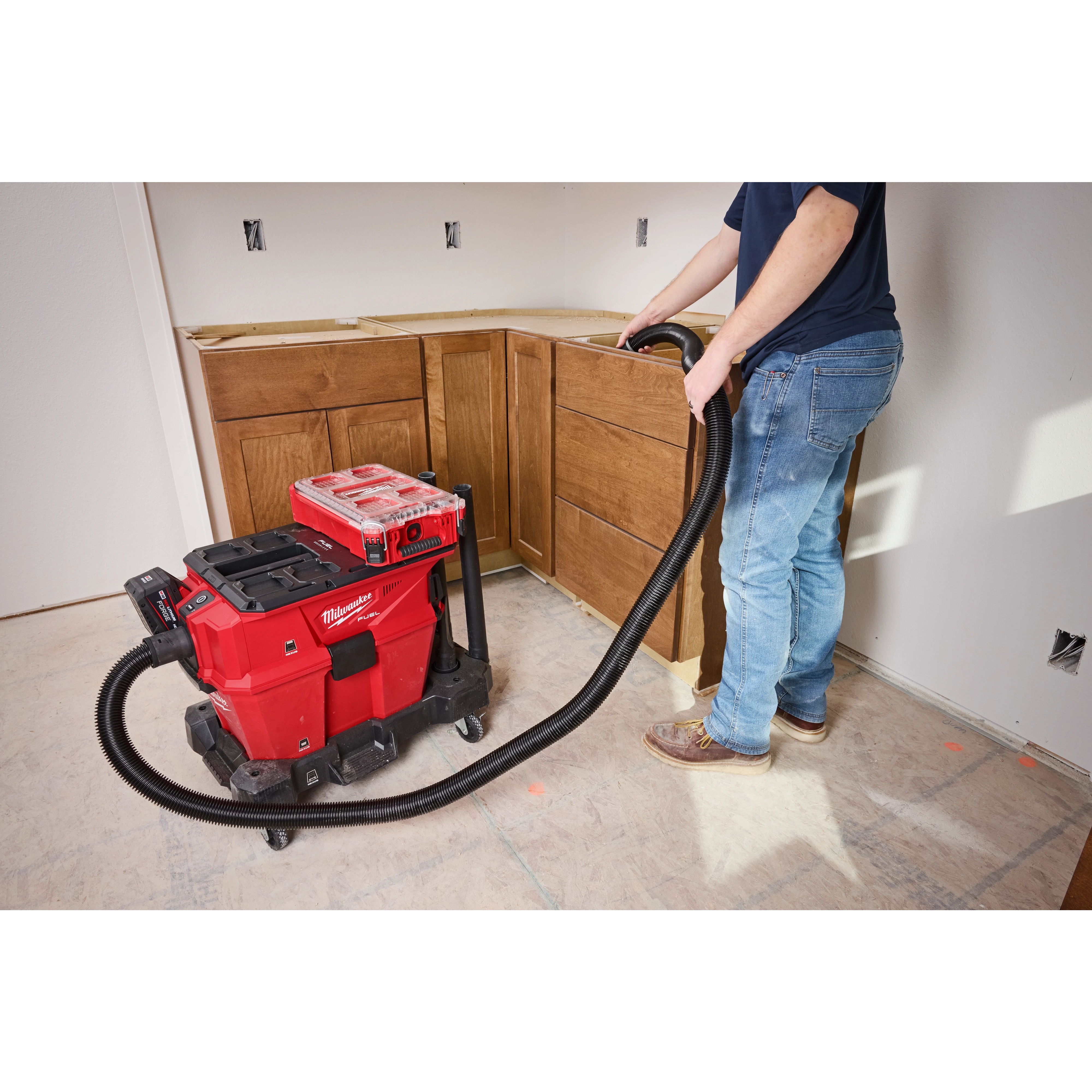 A person operates the M18™ FUEL™ NEXUS™ 6 Gallon Wet/Dry Vacuum with PACKOUT™ Compatibility by a wooden cabinet assembly. The red vacuum is on wheels and has a black hose attached to it. The vacuum is positioned on a tiled floor in a partially completed room.