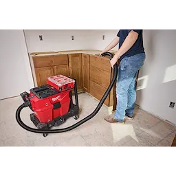 A person operates the M18™ FUEL™ NEXUS™ 6 Gallon Wet/Dry Vacuum with PACKOUT™ Compatibility by a wooden cabinet assembly. The red vacuum is on wheels and has a black hose attached to it. The vacuum is positioned on a tiled floor in a partially completed room.