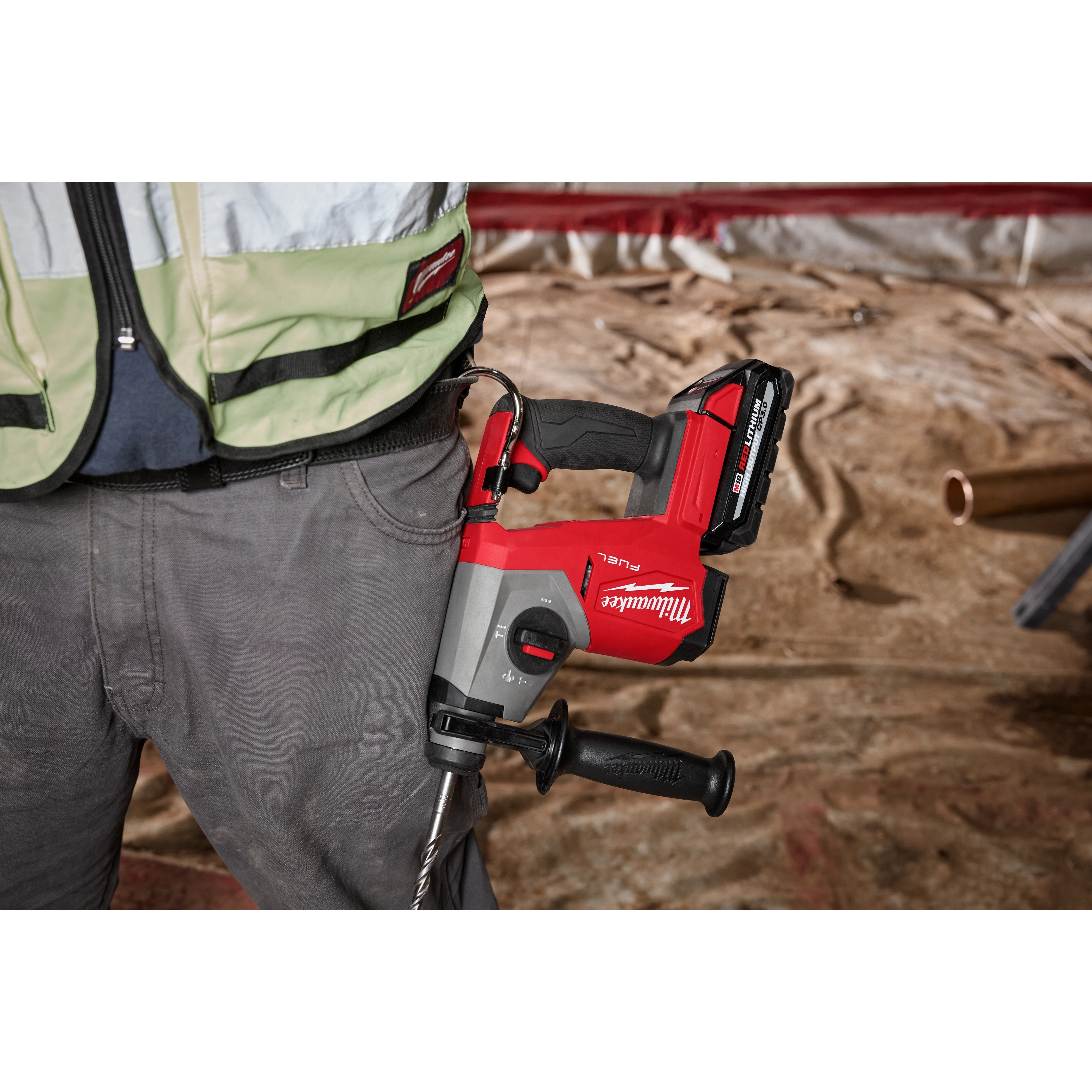 A red and black M18 FUEL™ 5/8" SDS Plus Rotary Hammer is attached to a worker's belt. The image showcases the power tool and the waist area of the worker. The background depicts a construction site.