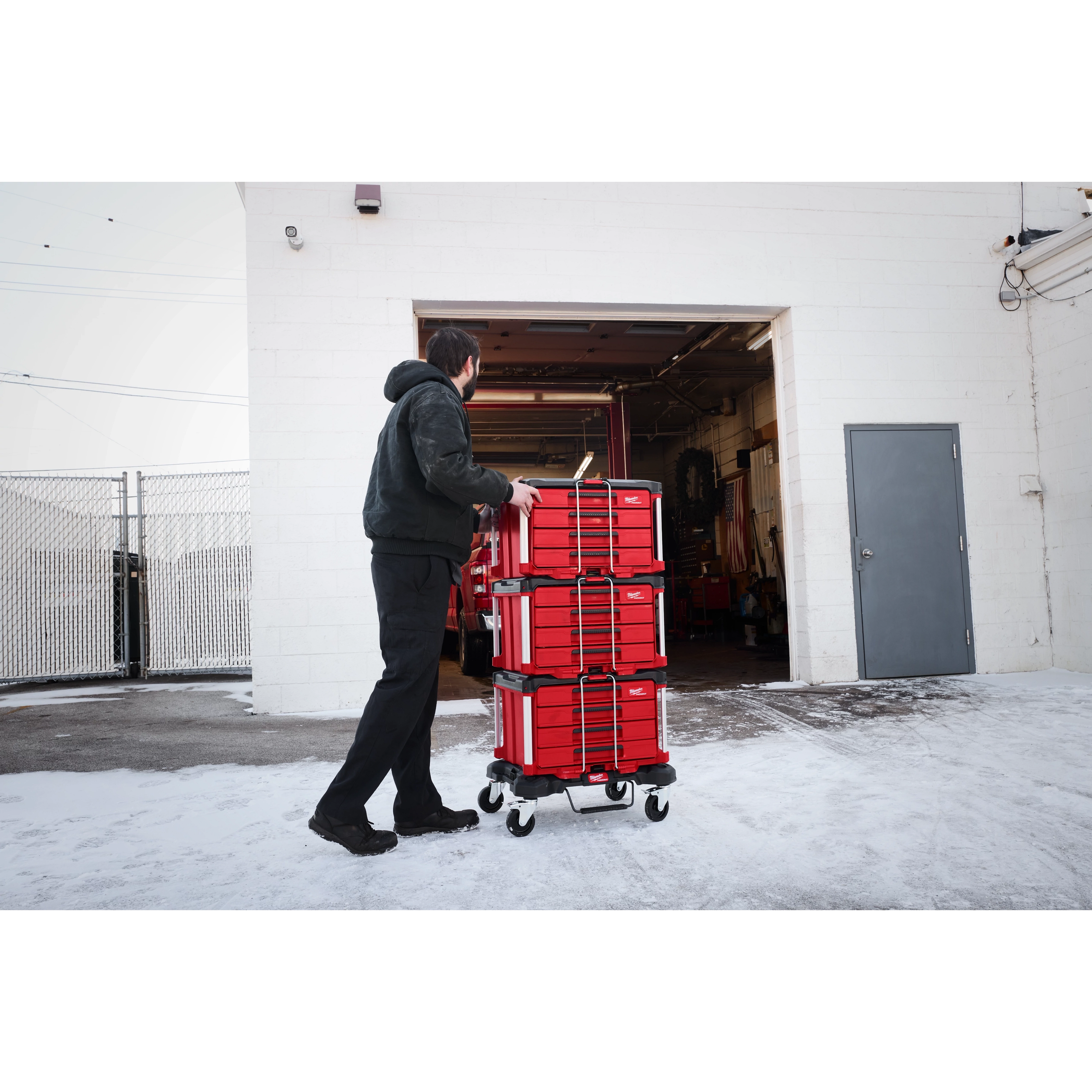 A person pushes a 366pc. Master Mechanics Hand Tool Set with PACKOUT™ Drawers and Dolly into a garage. The tool set is stacked in red, wheeled drawers, and the scene is outdoors with snow on the ground.