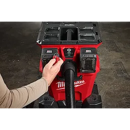 A person turns on the M18™ FUEL™ NEXUS™ Dual Battery Wet/Dry Vacuum Motor Head w/ PACKOUT™ Compatibility & VACLINK™. The red and black vacuum with two battery slots has a hose attached at the front, and the word "Milwaukee" is visible on the body.