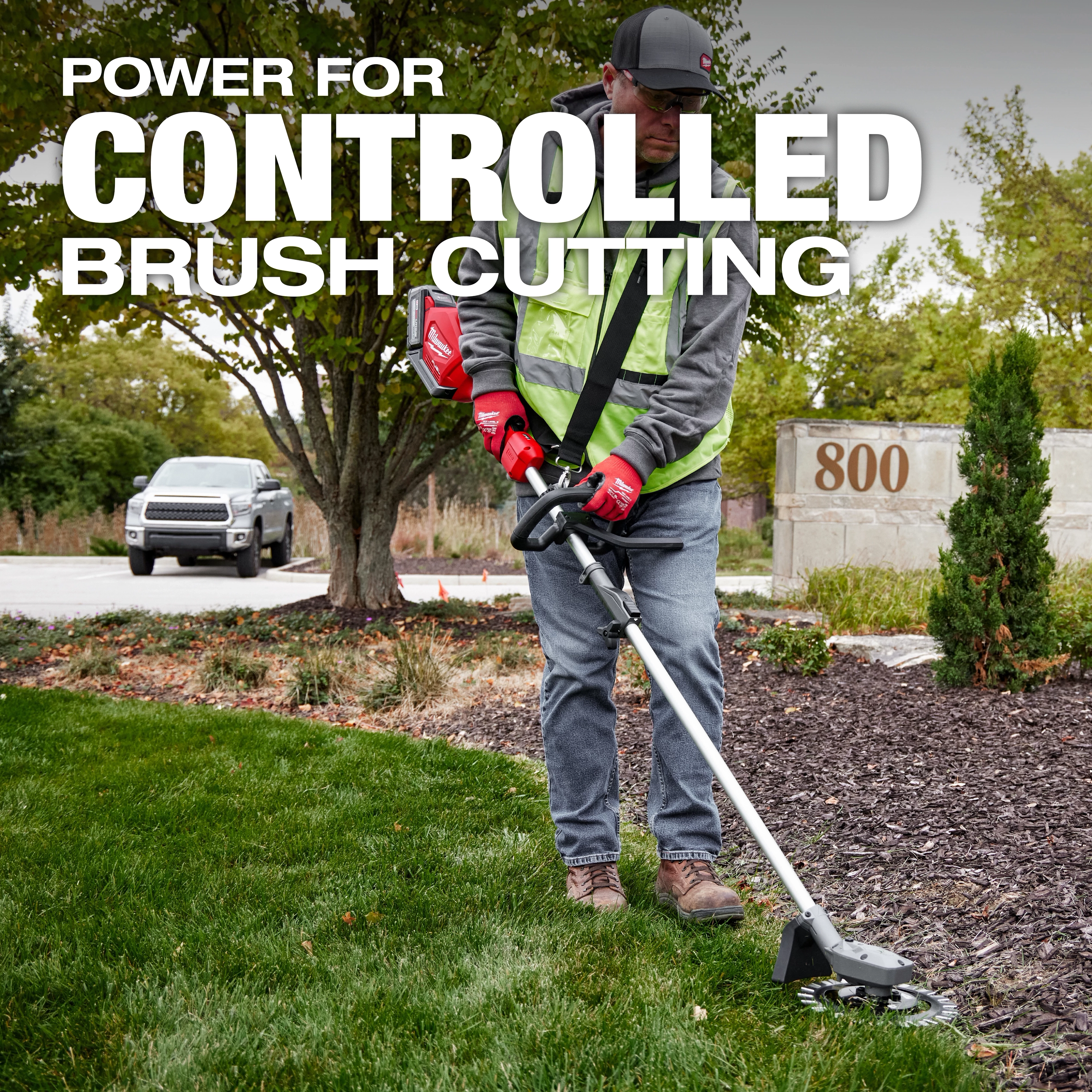 A person in a safety vest uses a brush cutter on a grassy and mulched area near a concrete sign marked "800." A white vehicle is parked in the background. The text overlay reads, "Power for Controlled Brush Cutting."