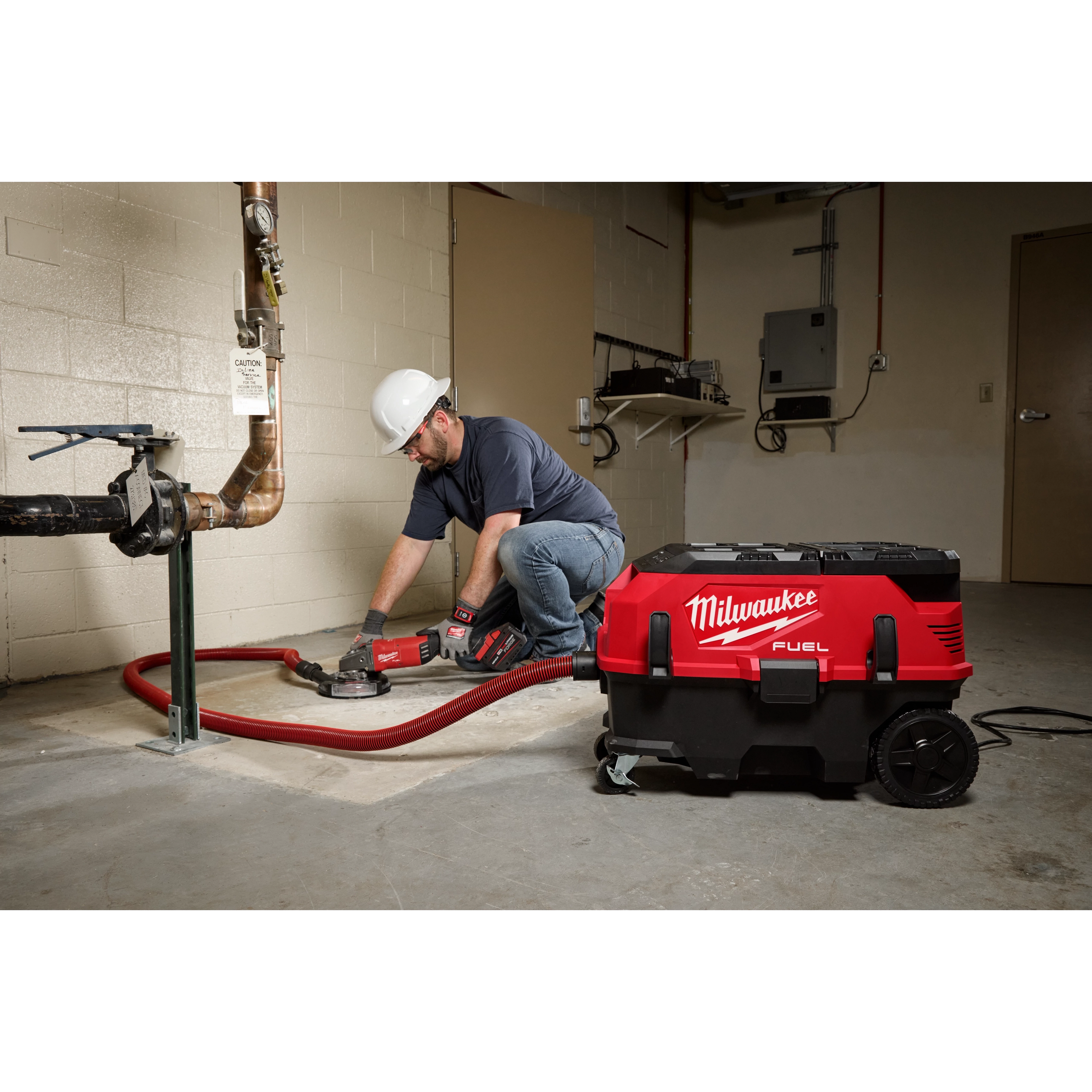 A person wearing a hard hat uses the M18 FUEL™ PACKOUT™ 9 Gallon Dual-Battery Dust Extractor with VACLINK™ in a workshop. The red and black dust extractor is connected to a tool via a red hose. The workplace has concrete flooring and visible pipes.