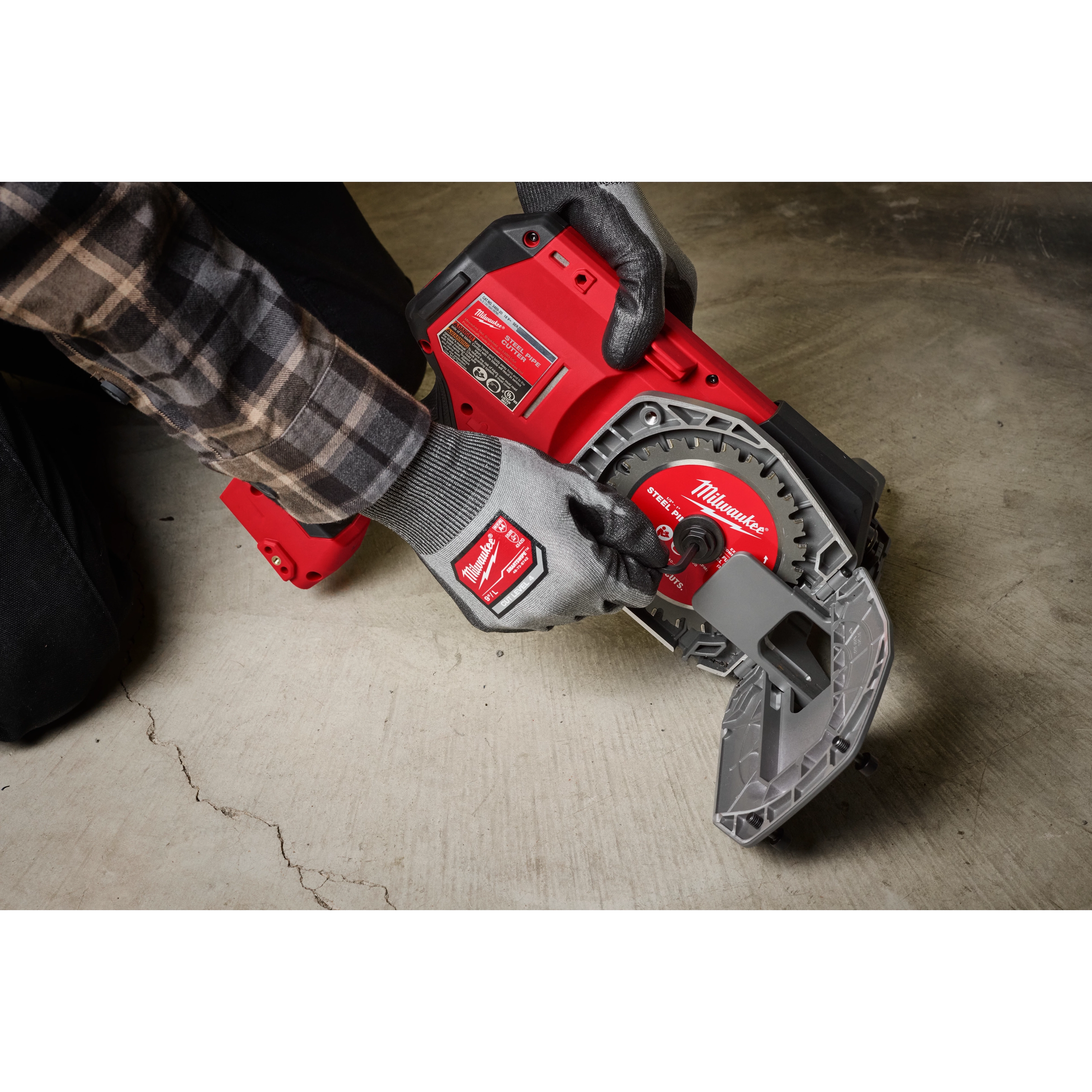 A person wearing work gloves operates a Milwaukee brand M18 FUEL™ ½”-1” Steel Pipe Cutter on a concrete floor. The tool is red and grey, showing the cutter's mechanisms and design features.