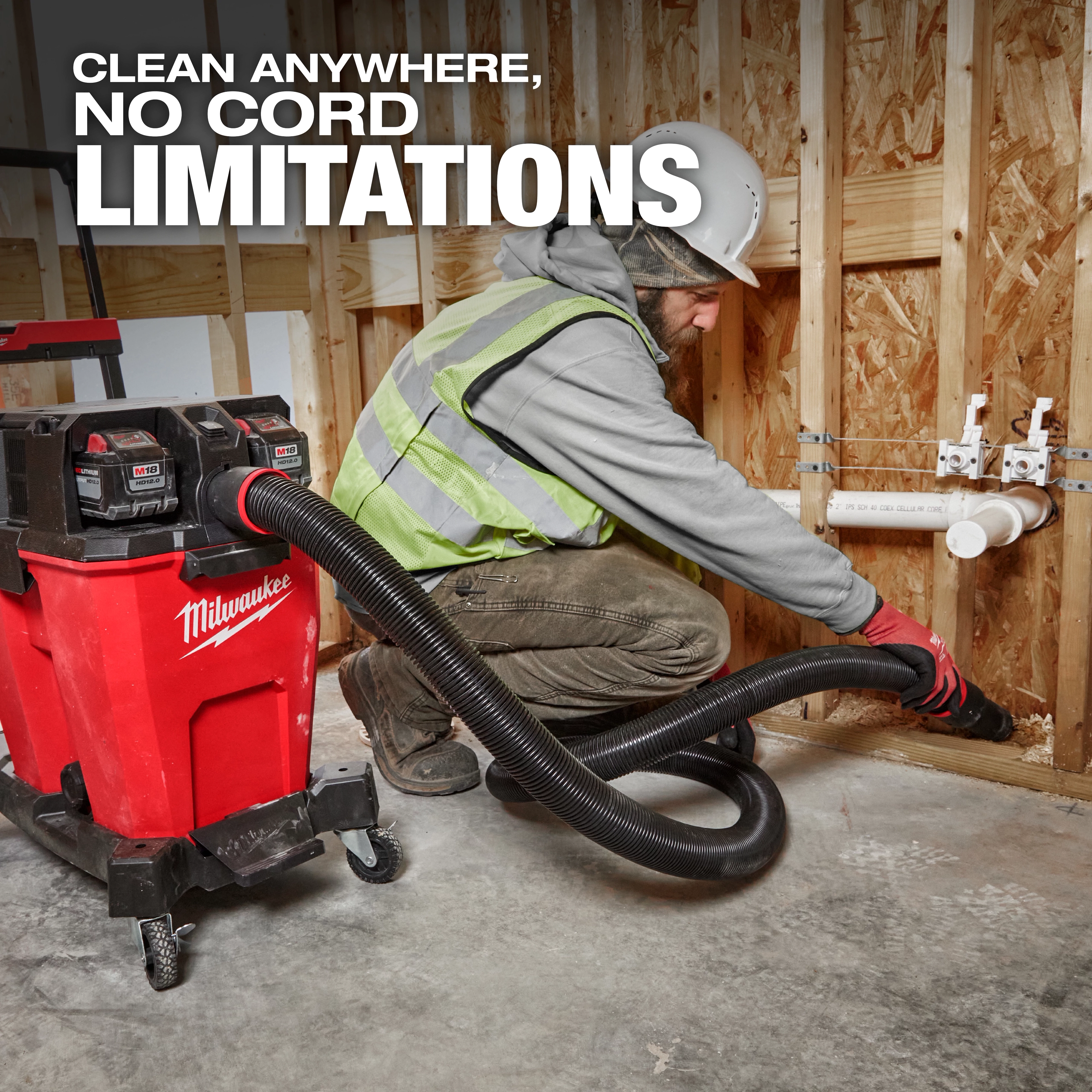 A worker uses the Milwaukee M18 FUEL™ NEXUS™ 12 Gallon Dual-Battery Wet/Dry Vacuum Kit to clean debris. The vacuum is cordless and positioned on a construction site with wooden framing and plumbing pipes in the background.