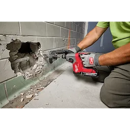 A person in work gloves uses the M18 FUEL™ 5/8" SDS Plus Rotary Hammer with Dust Extractor Kit to create a hole in a tiled wall, revealing pipes. The tool features a HAMMERVAC™ 5/8" Dedicated Dust Extractor for dust collection.