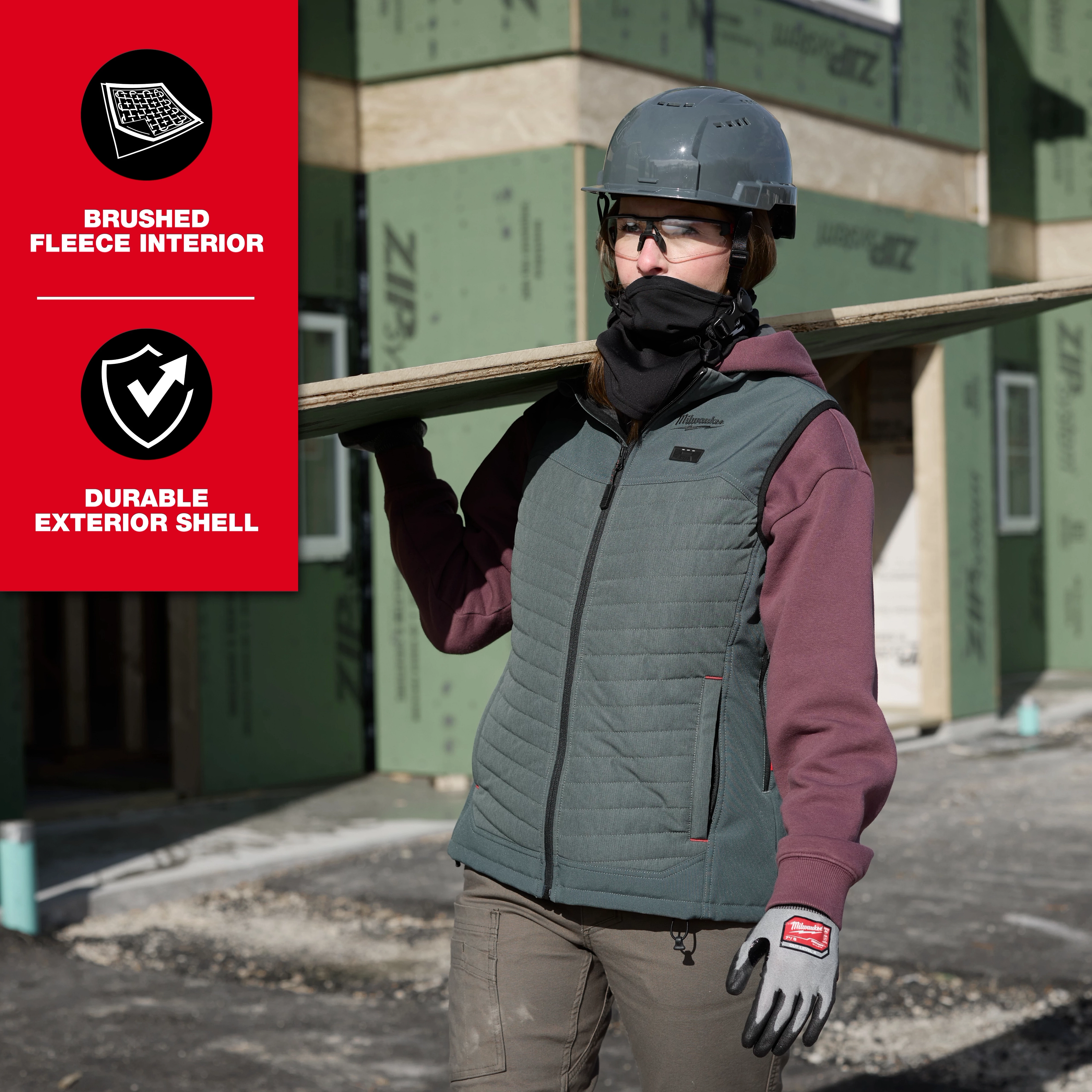 The WORKSKIN™ Balaclava is worn by a person at a construction site. They are also wearing a grey helmet, a green and black vest over a maroon top, beige pants, and gloves. The image highlights the balaclava's brushed fleece interior and durable exterior shell features.