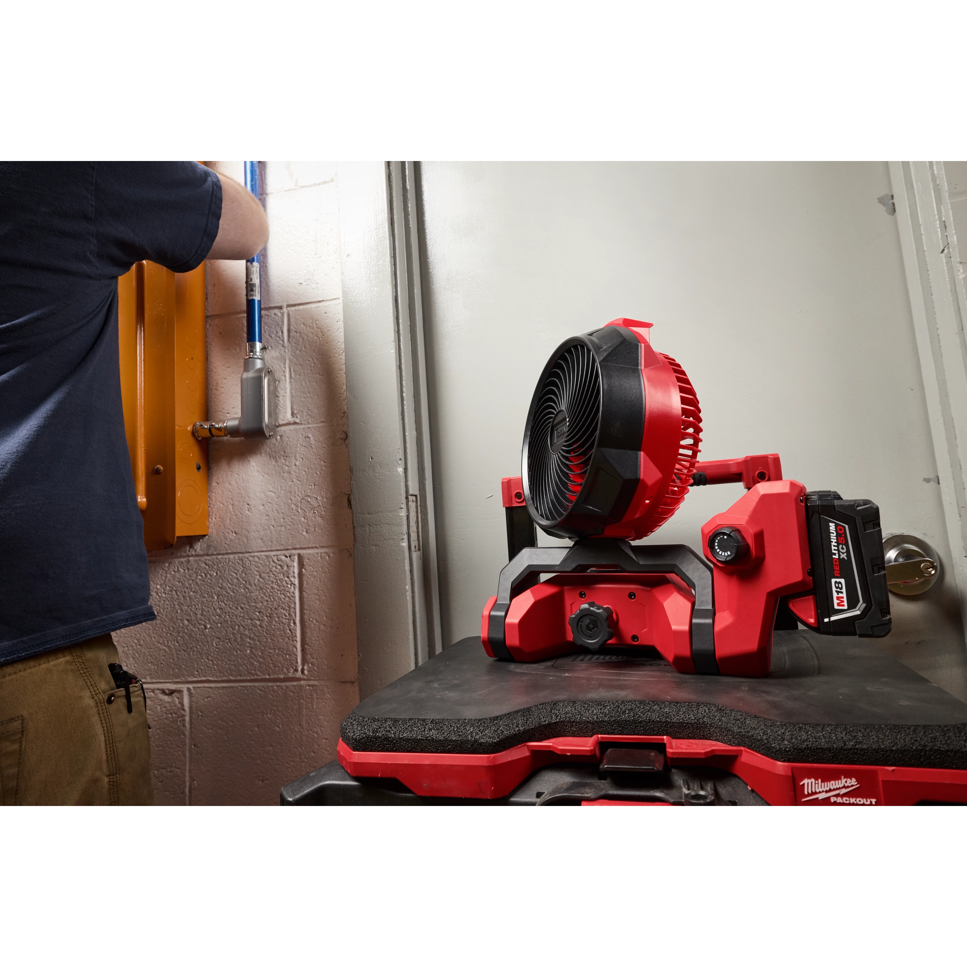 M18 Brushless Scissor Lift Fan in a workshop, placed on a red and black surface, with a person working in the background.
