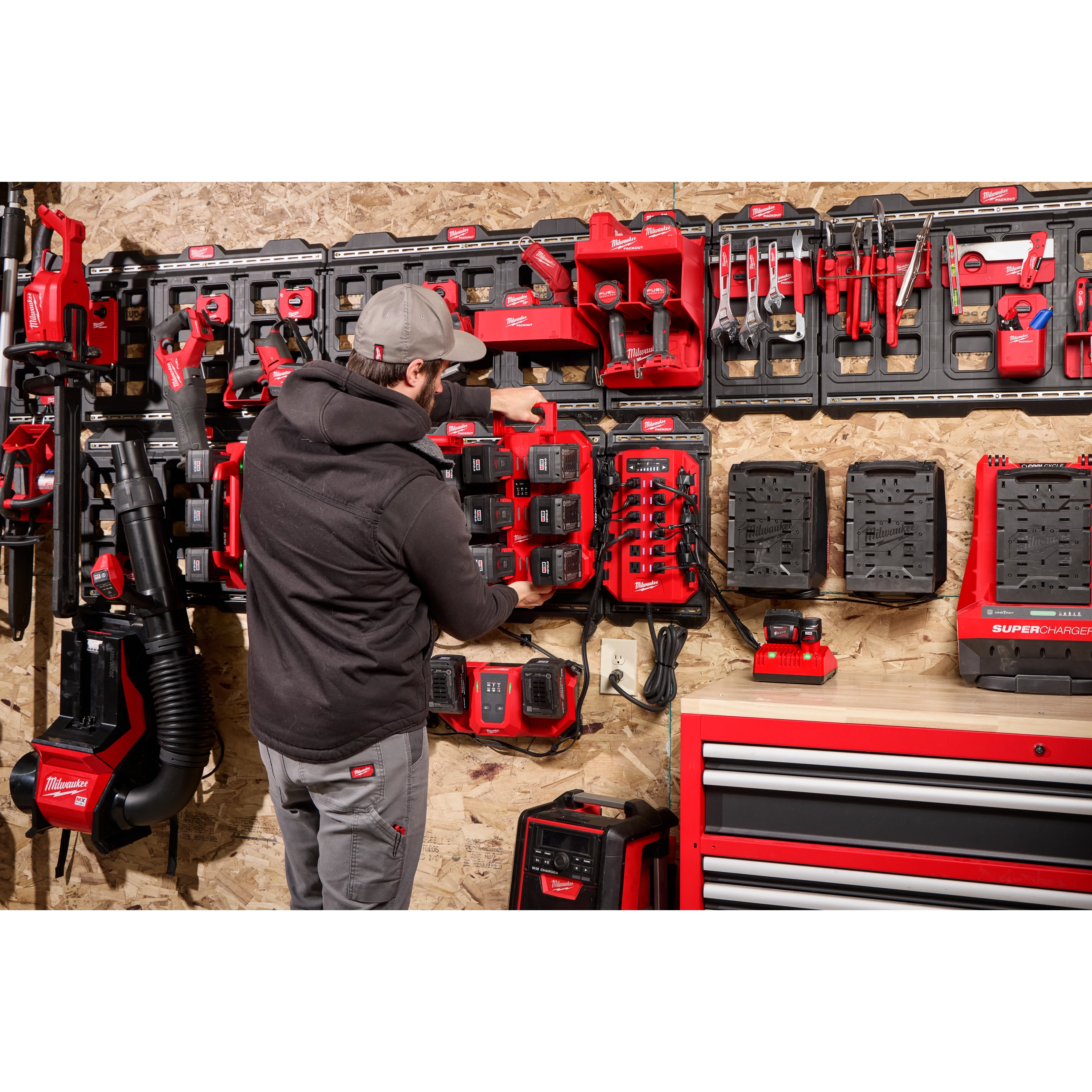 A person in a black hoodie and gray cap stands in a workshop, organizing red and black tools on a pegboard. The foreground includes labeled items, particularly a "Power Manager – 20 Amp Circuit" device.