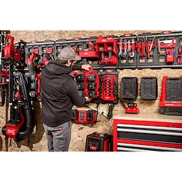 A person in a black hoodie and gray cap stands in a workshop, organizing red and black tools on a pegboard. The foreground includes labeled items, particularly a "Power Manager – 20 Amp Circuit" device.