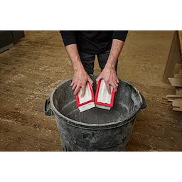 A person is holding two rectangular HEPA filters with red borders above a large trash bin. The scene appears to be in a workshop with a wooden floor. The image depicts a HEPA%20Filter%20%282PK%29.