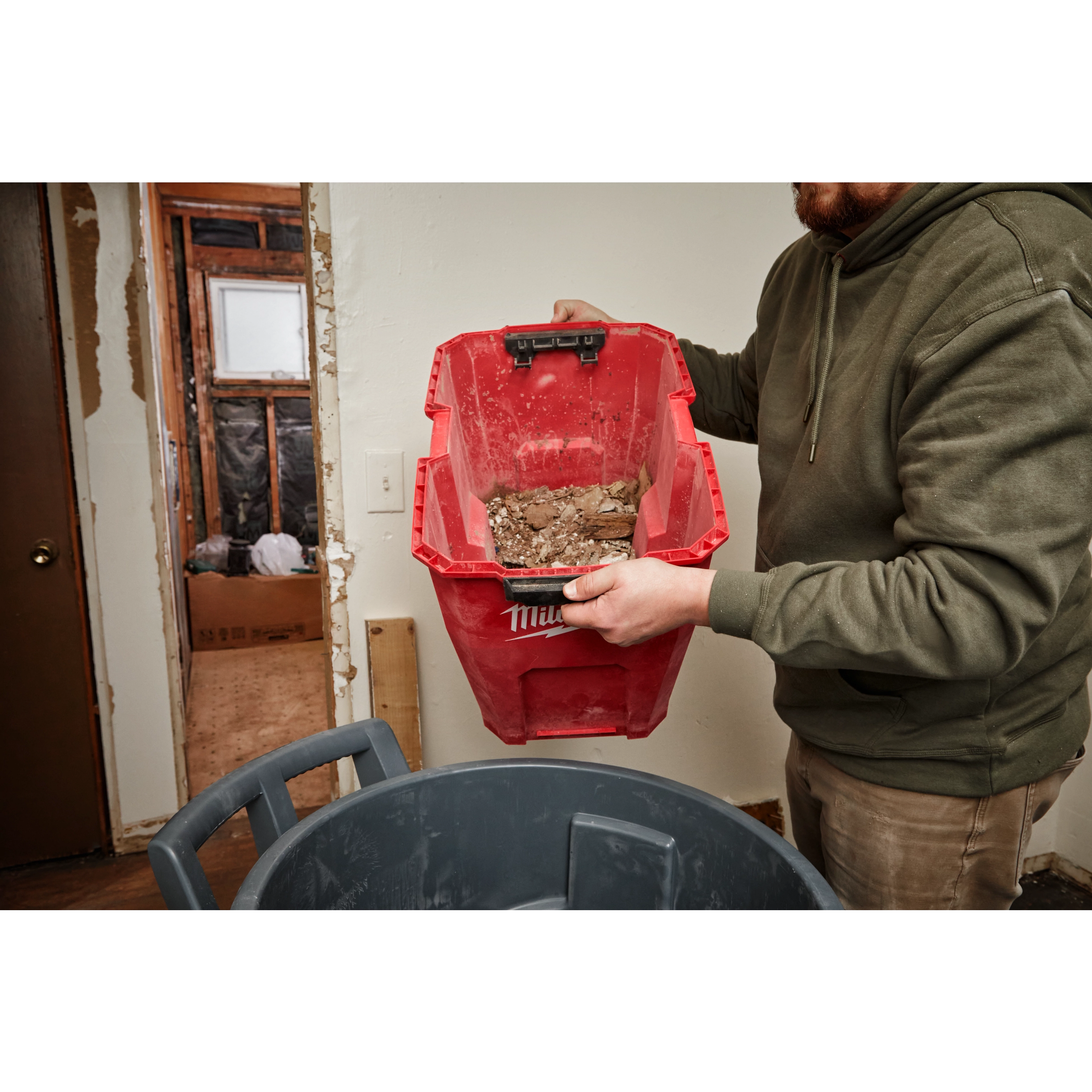 Person holding a red 12 Gallon Wet/Dry Vacuum Tank and emptying its contents into a gray trash bin in a room under renovation.