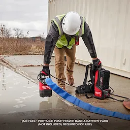 A person wearing a hard hat and safety vest operates an MX FUEL™ 1HP 2" Submersible Pump next to a shallow puddle. The pump is connected to a blue hose and a red and black power base. The text indicates a portable pump power base and battery pack are required but not included.
