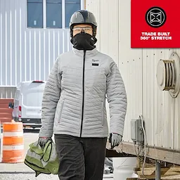 A person wearing a grey WORKSKIN™ Balaclava, a light grey jacket, and grey gloves is holding a green hard hat while standing outdoors near a building and safety cones. The image highlights the product's 360° stretch feature with a "TRADE BUILT 360° STRETCH" tag in the top right corner.
