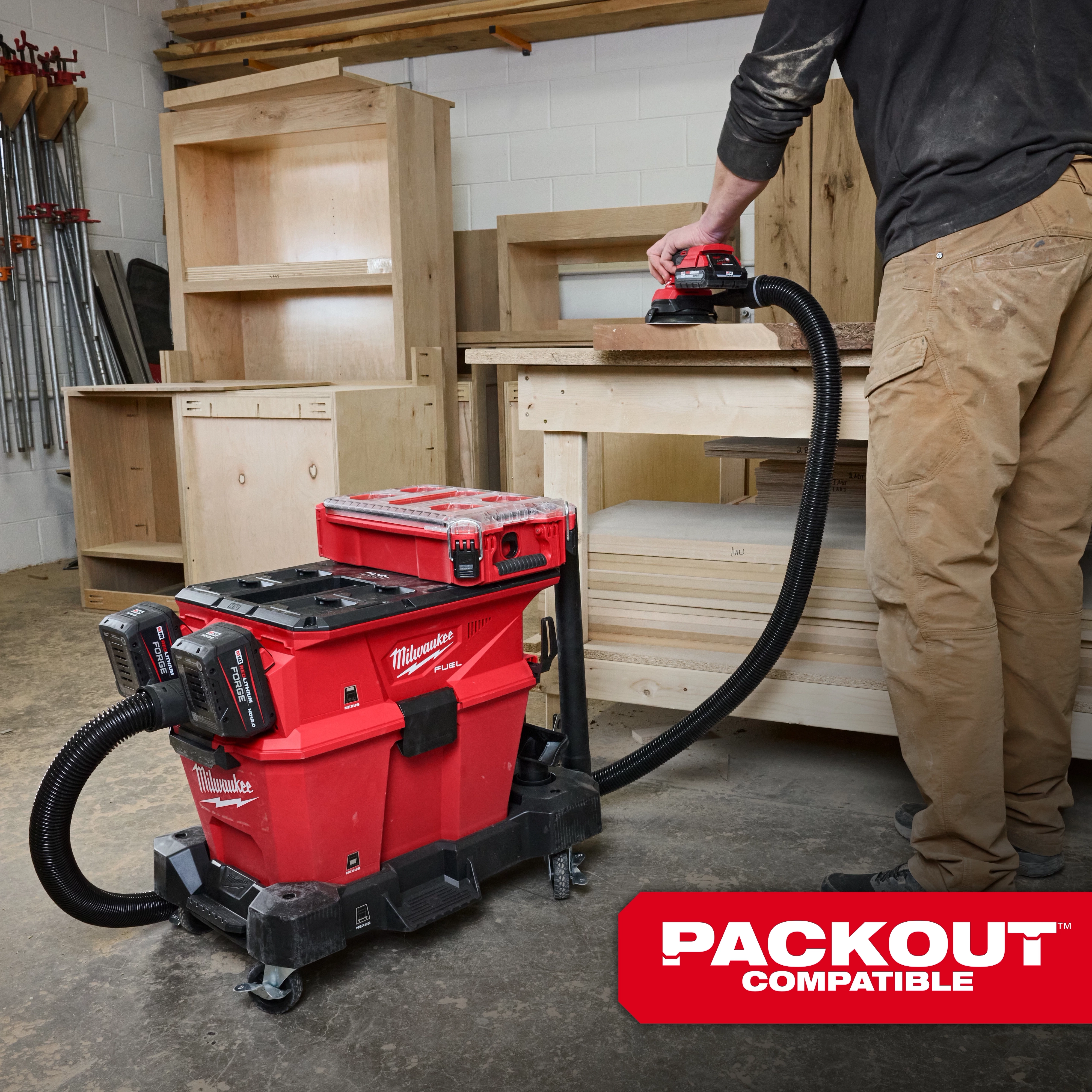 The M18 FUEL™ NEXUS™ Dual Battery Wet/Dry Vacuum Motor Head with PACKOUT™ Compatibility & VACLINK™ is shown in a workshop. It is a red portable vacuum cleaner on wheels, connected to a hose, and features two battery packs for power. A person operates the vacuum on a wooden surface.
