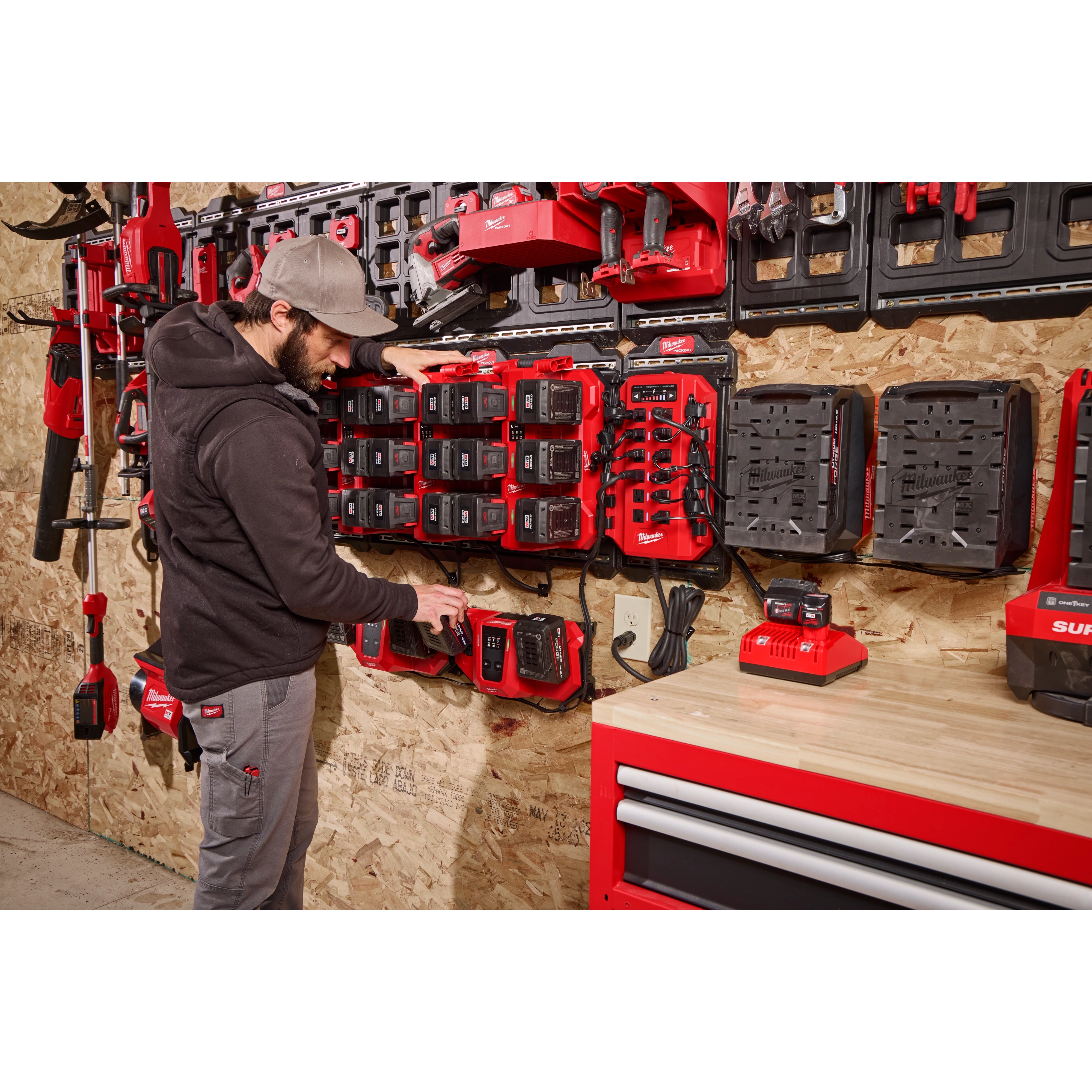 A person stands near a wall-mounted charging station, organizing tools and batteries labeled Power Manager – 20 Amp Circuit. The setup includes various power tools, chargers, and a red tool chest with drawers.