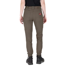 A person wearing Women's GRIDIRON™ Double Knee Jogger Pants - Brown. The brown jogger pants feature multiple pockets, a red logo patch on the back pocket, and elastic cuffs at the ankles. The person is shown from the back, wearing black boots.