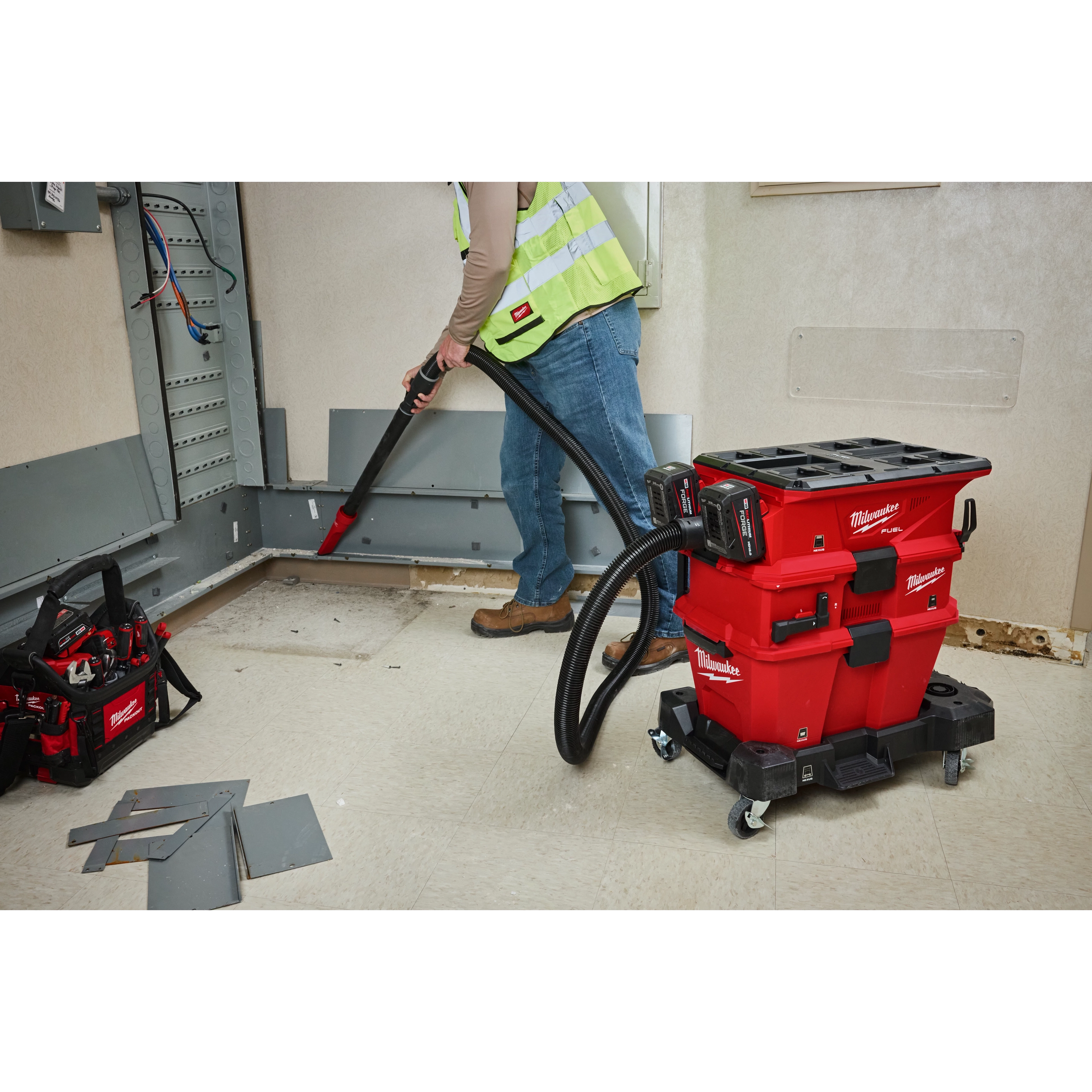 A worker uses the M18™ FUEL™ NEXUS™ Dual Battery Wet/Dry Vacuum Motor Head with PACKOUT™ Compatibility & VACLINK™ to clean up a work area. The red vacuum with black hoses and attachments is positioned on a wheeled base for mobility. Nearby, a tool bag and debris are visible on the floor.