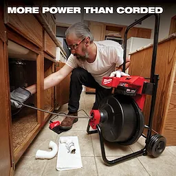 The Milwaukee MX FUEL Drum Machine is shown in a kitchen, being used to unclog pipes under a sink. A person operates the device, which features a large drum and handles, with plumbing components scattered nearby on the tiled floor. Text above reads, "More power than corded."