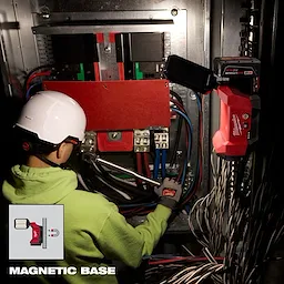 A technician works on electrical cables using a wrench. Nearby, the M12™ ROVER™ Multi-Directional Flood Light w/ PACKOUT™ Compatibility is magnetically attached to the panel, illuminating the work area. An inset image shows its magnetic base. The background includes various cables and electrical components.