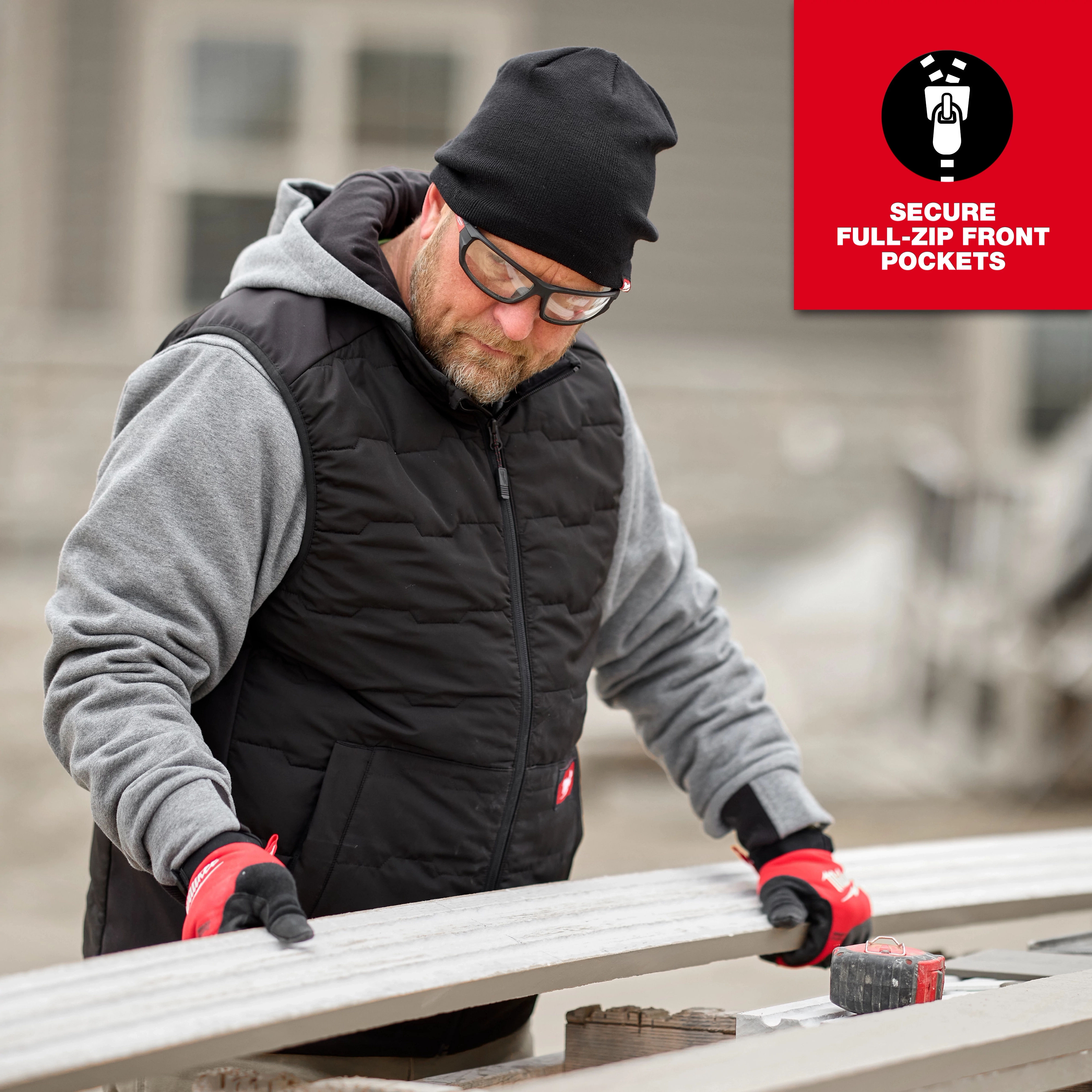 A person is wearing a Milwaukee M12 Heated AXIS Vest in black, with a grey hoodie underneath and red work gloves. The product features secure full-zip front pockets. They appear to be working with tools and materials at an outdoor construction site.