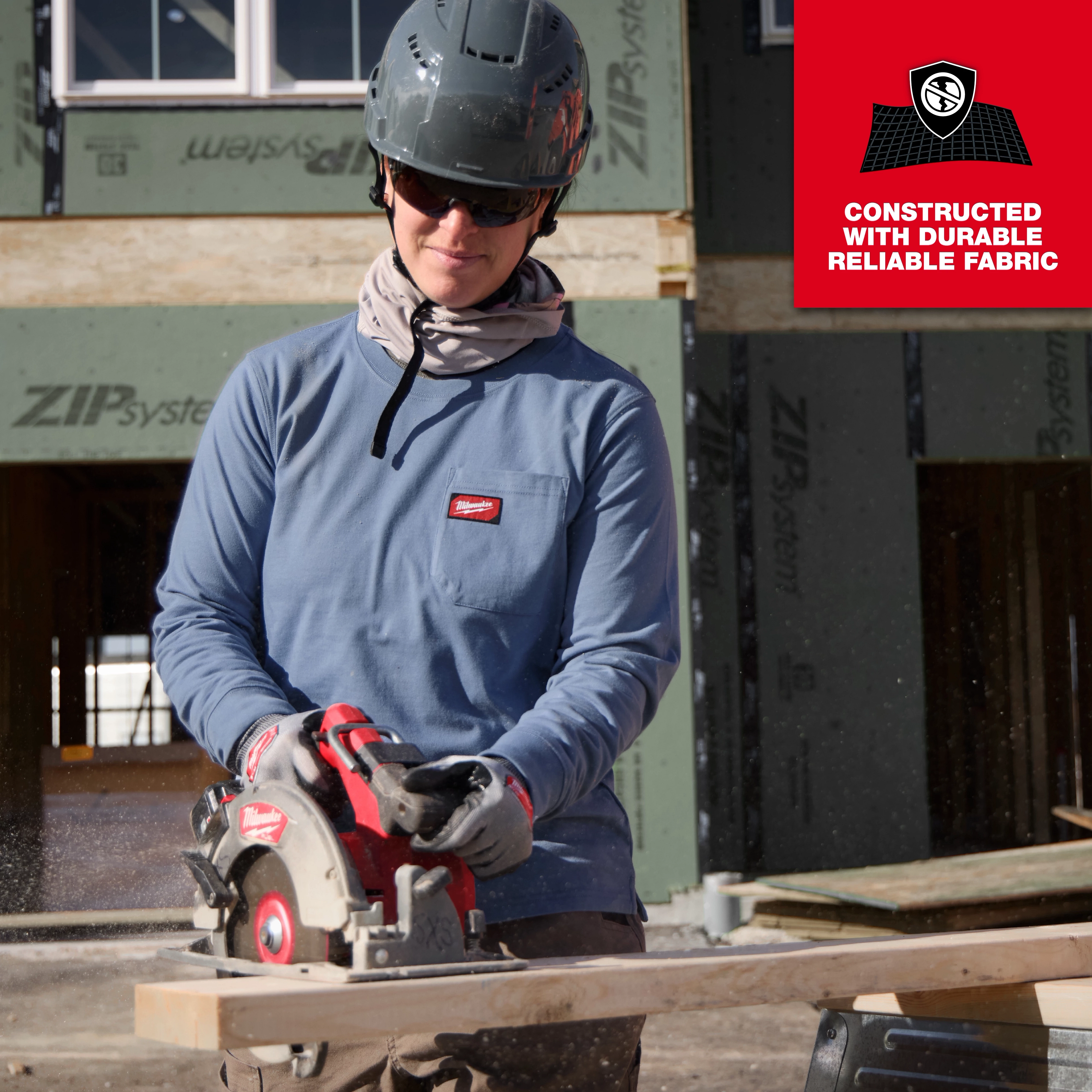 A worker wearing a Women's GRIDIRON™ Pocket T-Shirt - Long Sleeve in blue uses a circular saw on a wooden plank at a construction site. A red label indicates the shirt is constructed with durable, reliable fabric.