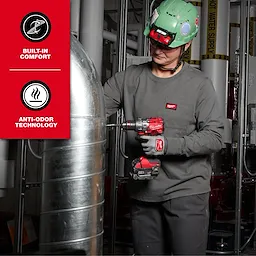 A person uses a red cordless power tool near metal pipes wearing the Women’s GRIDIRON™ Pocket T-Shirt - Long Sleeve. The shirt is dark grey with a pocket. The image highlights built-in comfort and anti-odor technology features of the shirt.