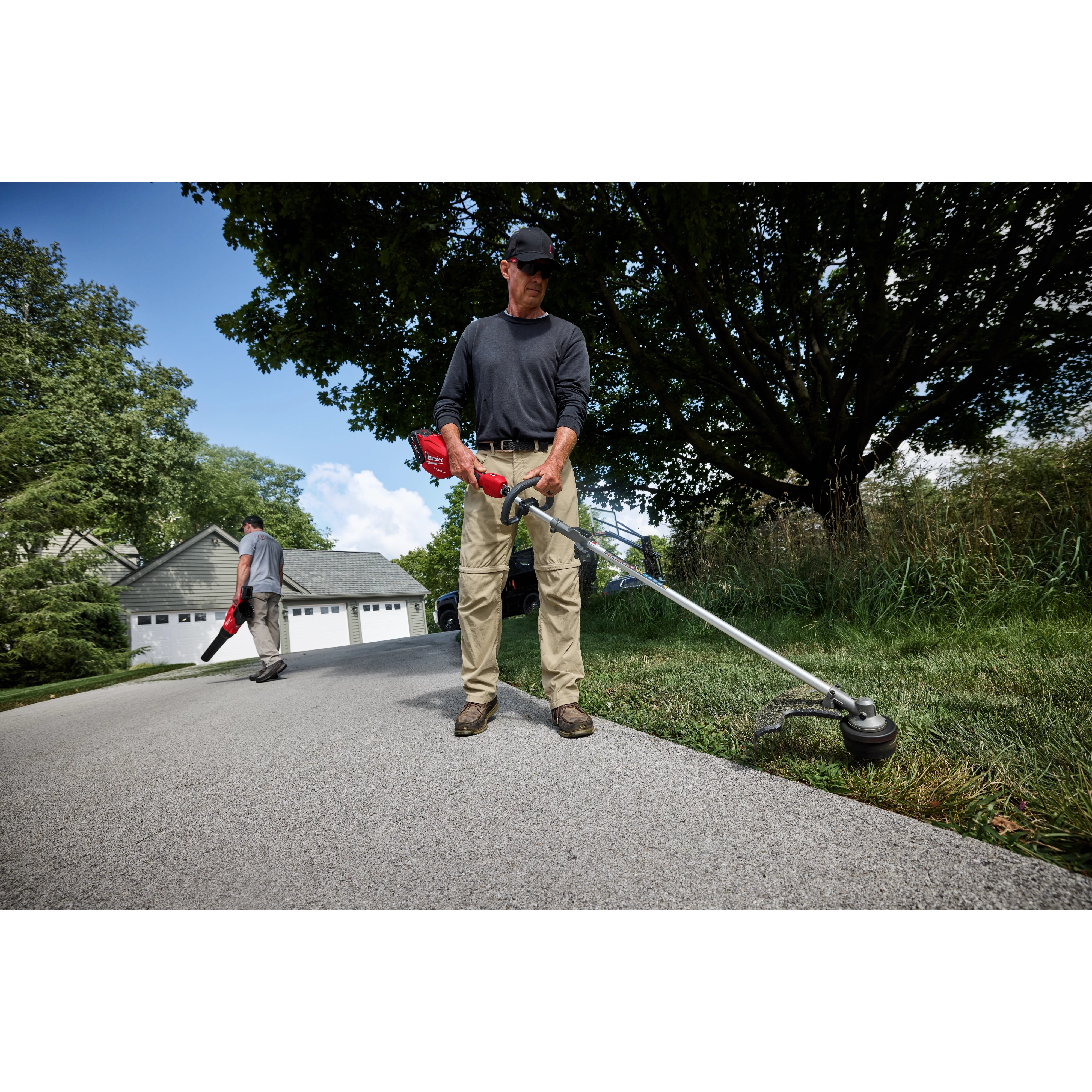 A person uses the M18 FUEL™ Power Head w/ QUIK-LOK™ to trim grass along a driveway. Another person in the background uses a blower. They are working near a house with a garage and trees around.