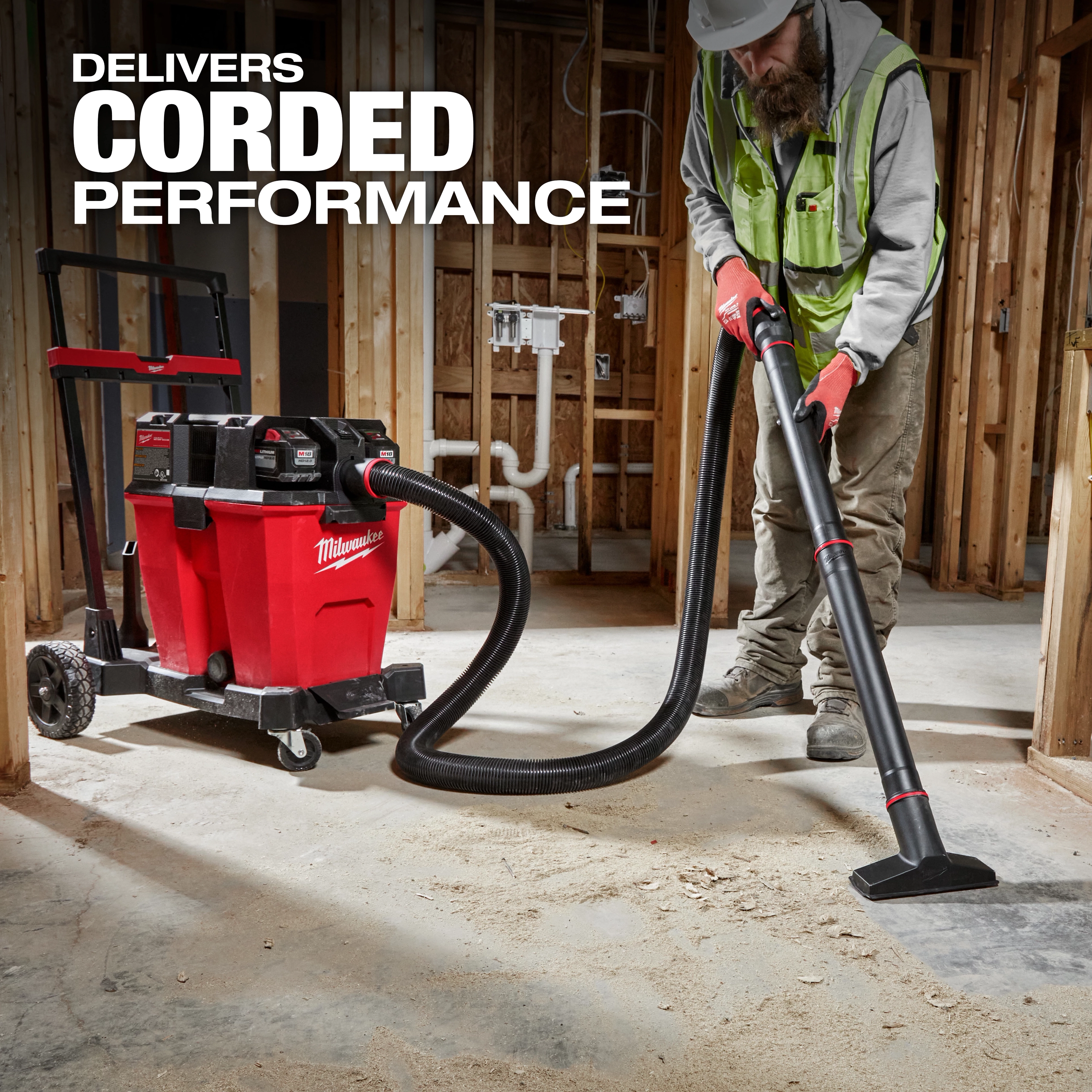 A worker uses the M18 FUEL™ NEXUS™ 12 Gallon Dual-Battery Wet/Dry Vacuum Kit to clean up construction debris. The red and black vacuum unit is equipped with wheels, a hose, and multiple battery slots. Text reads "Delivers Corded Performance." The background features an unfinished building interior.
