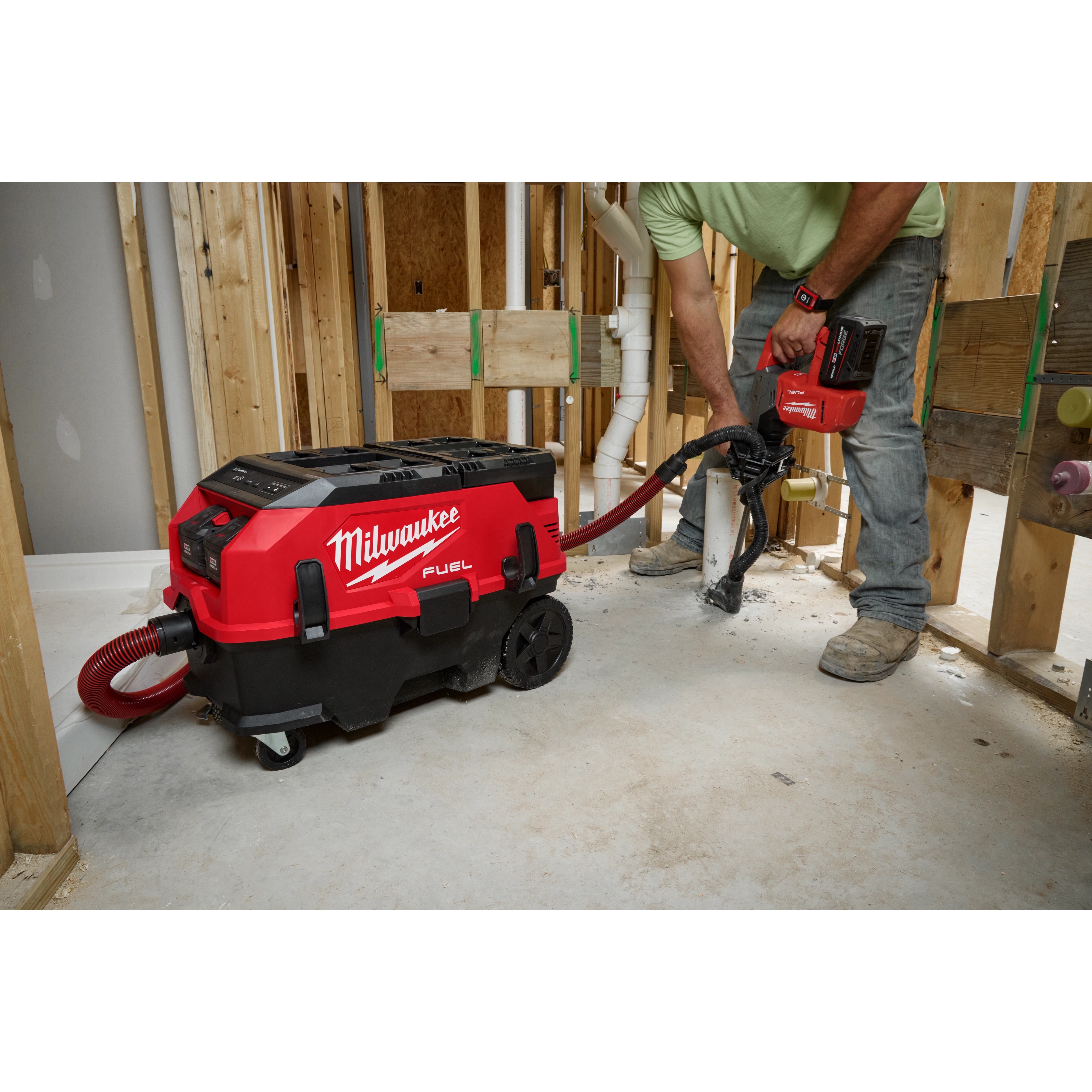 A person operates a red M18 FUEL™ PACKOUT™ 9 Gallon Dual-Battery Dust Extractor w/ VACLINK™ in a construction setting. The dust extractor is connected to a cutting tool, collecting dust and debris. The person is standing beside wooden studs and plumbing pipes in an unfinished room.