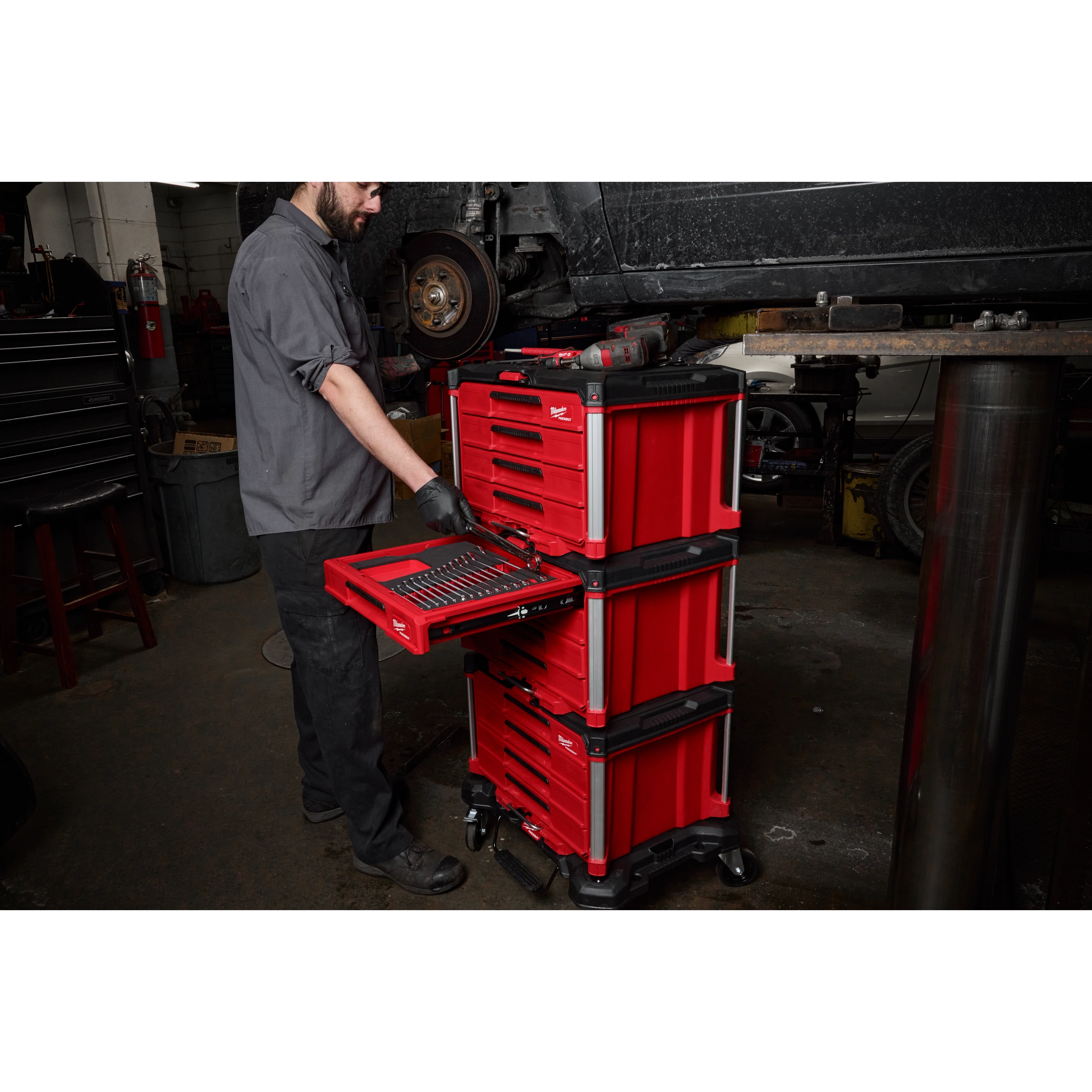 A mechanic is using a 366pc. Master Mechanics Hand Tool Set with PACKOUT™ Drawers and Dolly in a garage. The red tool chest is on wheels and has multiple drawers filled with tools.